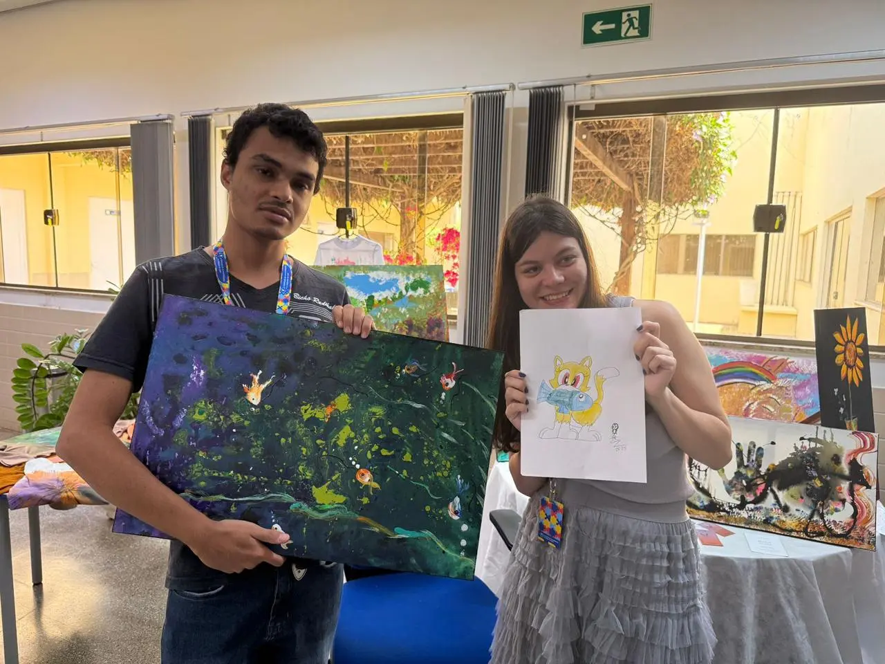 Júlio e Maria Clara exibem orgulhosos suas criações: uma pintura colorida e um desenho animado. O ambiente expositivo, repleto de quadros e luz natural, valoriza a inclusão e a arte como forma de comunicação.