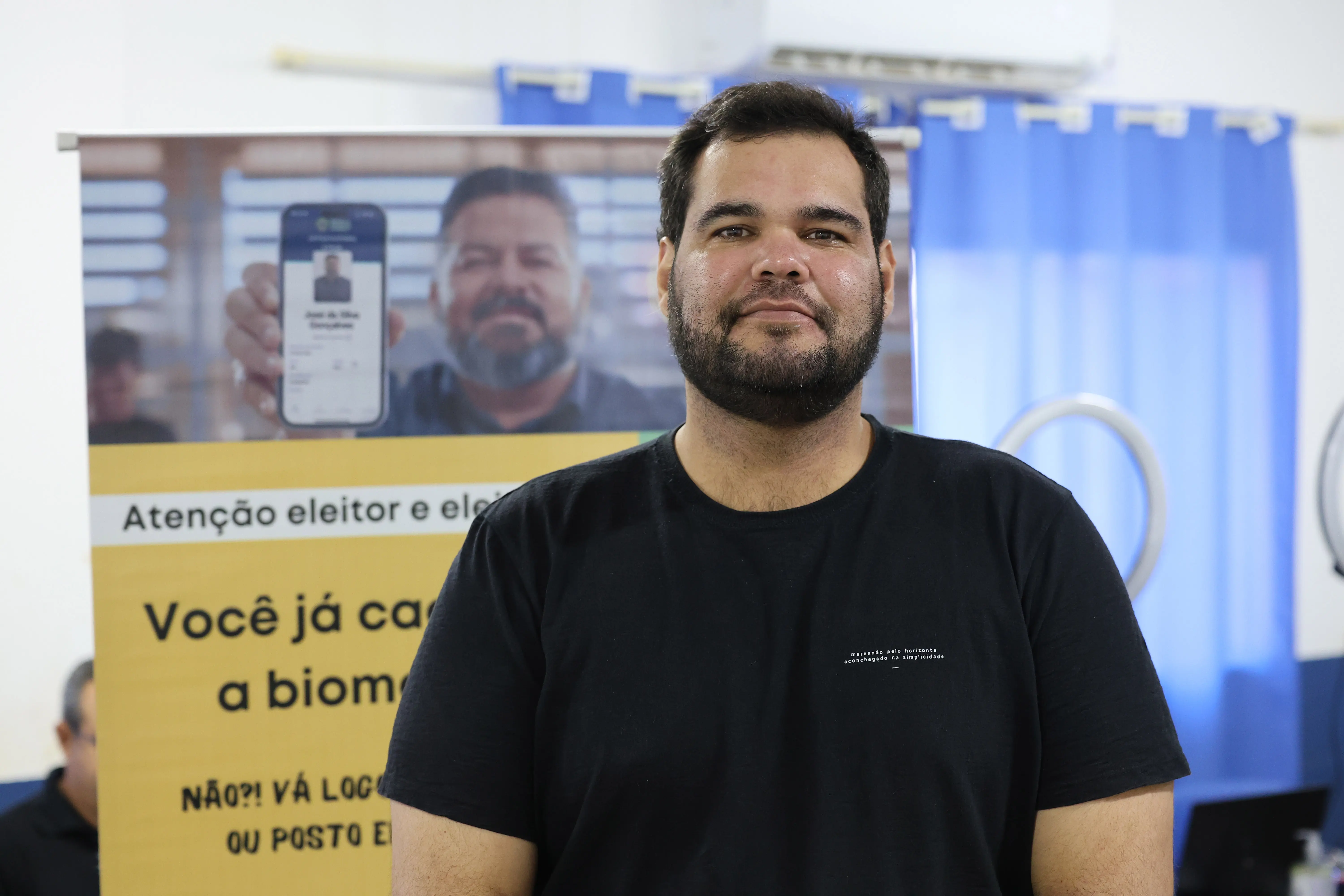 Homem de barba e camiseta preta sorri levemente diante de cartaz do Tribunal Regional Eleitoral sobre cadastramento biométrico. Atrás dele, cortinas azuis e equipamentos de registro completam o ambiente.