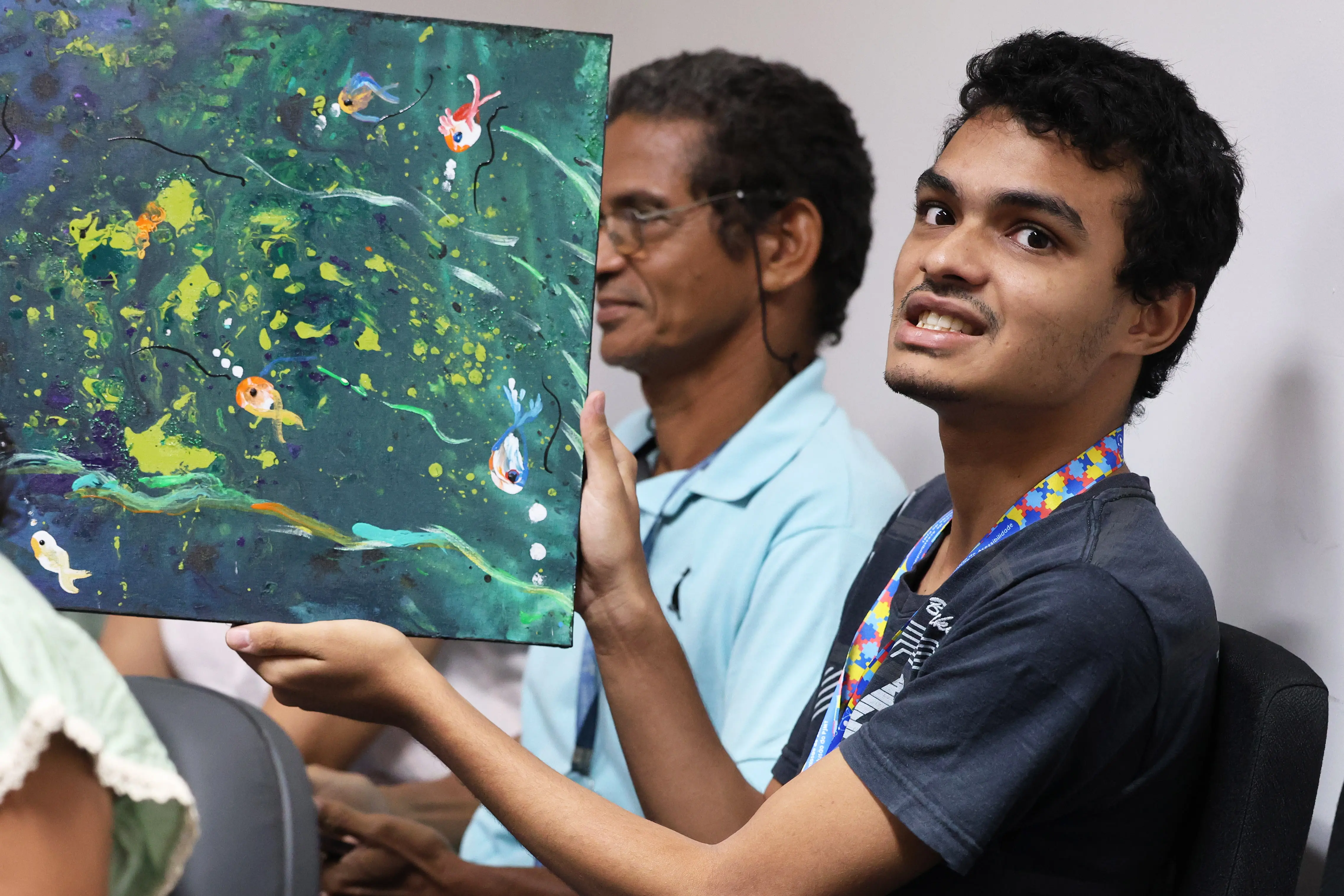 Júlio mostra sua pintura com peixes coloridos em tons de verde e azul, sorrindo levemente. Ao fundo, pessoas observam com admiração em sala iluminada, marcada por sensibilidade e incentivo à arte.