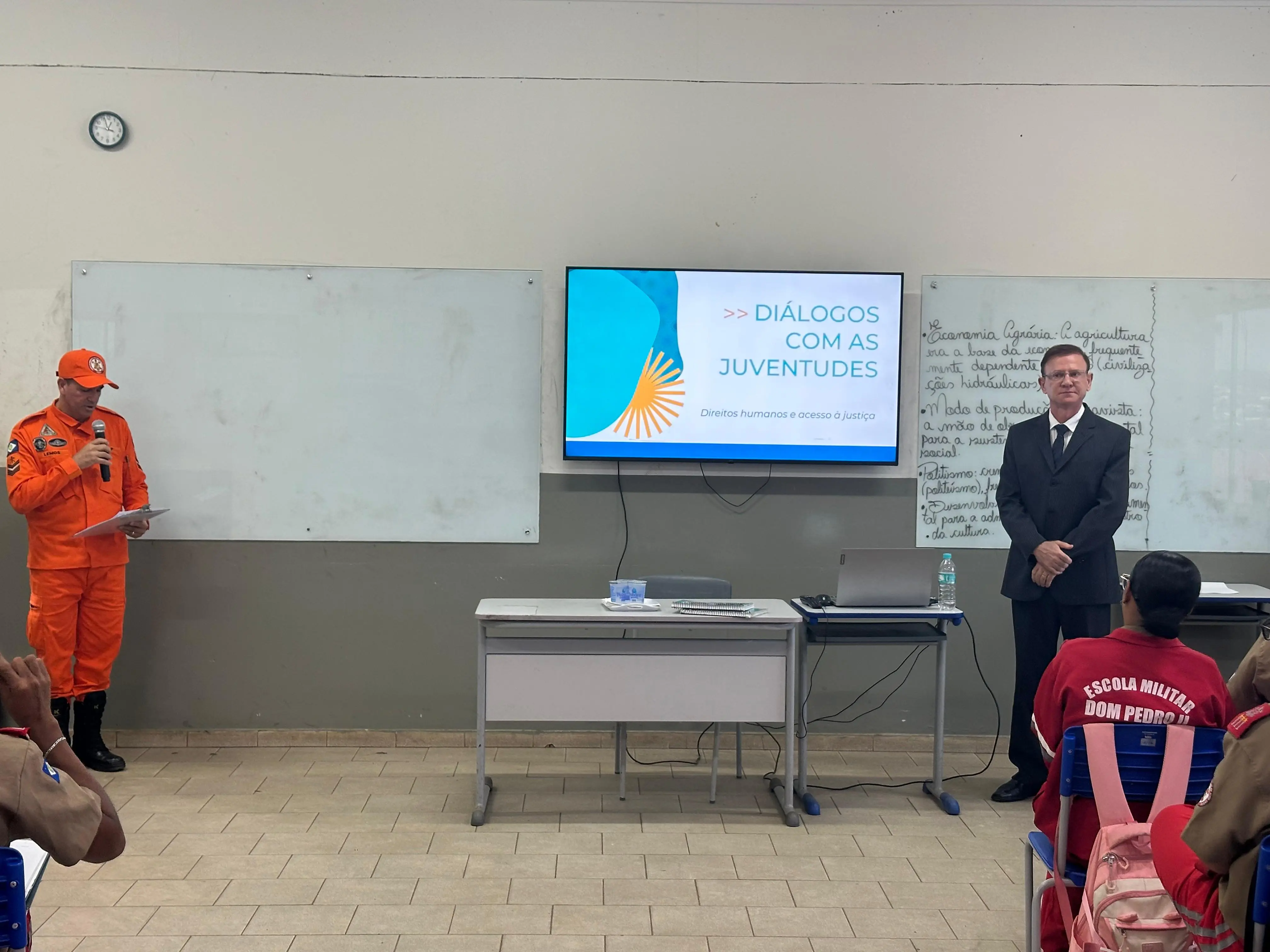 Juiz, de terno preto, é apresentado por bombeiro militar fardado de laranja durante palestra do projeto “Diálogos com as Juventudes”. A atividade ocorre em sala de aula iluminada, com quadro branco e televisão.