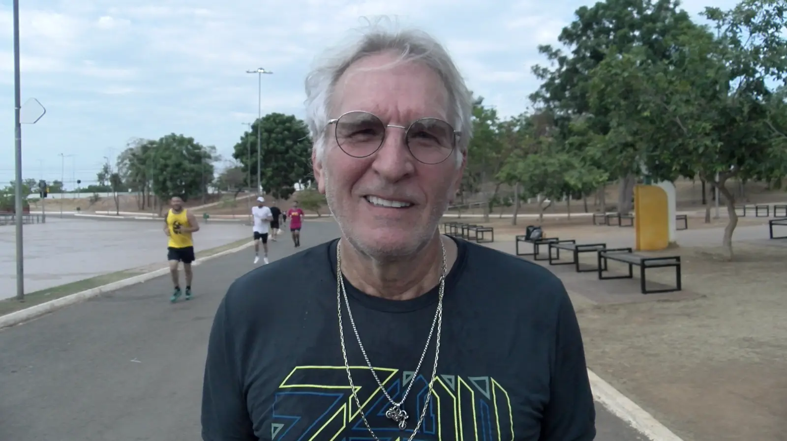 Homem de cabelos grisalhos e óculos sorri durante corrida em parque arborizado, usando camiseta escura e colares prateados.