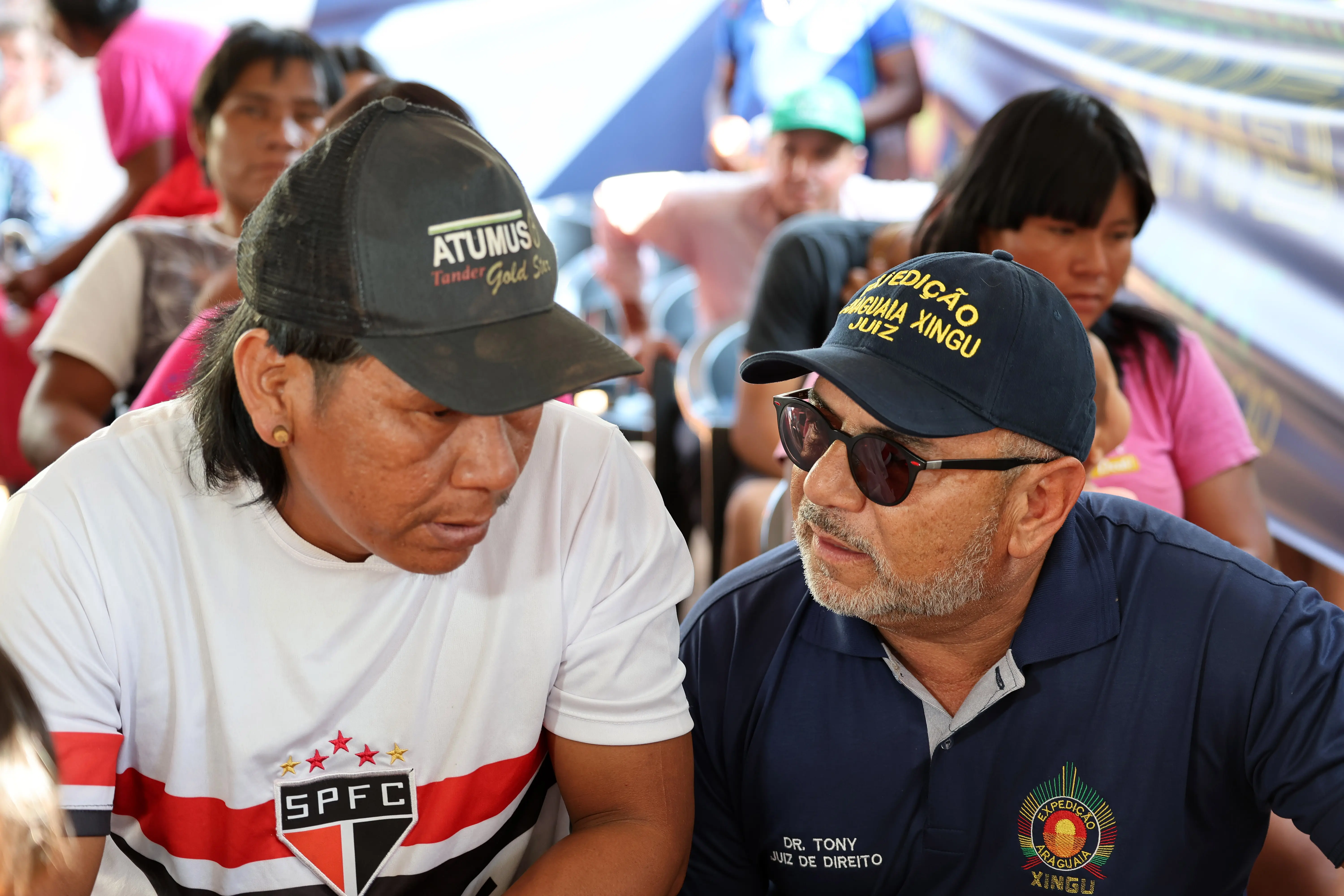 Homem indígena de pele morena, boné preto e camiseta branca do São Paulo Futebol Clube conversa atentamente com juiz de direito, que usa boné azul escuro, óculos e camiseta da Expedição Araguaia Xingu. Estão sentados entre várias pessoas em um espaço coberto e iluminado.