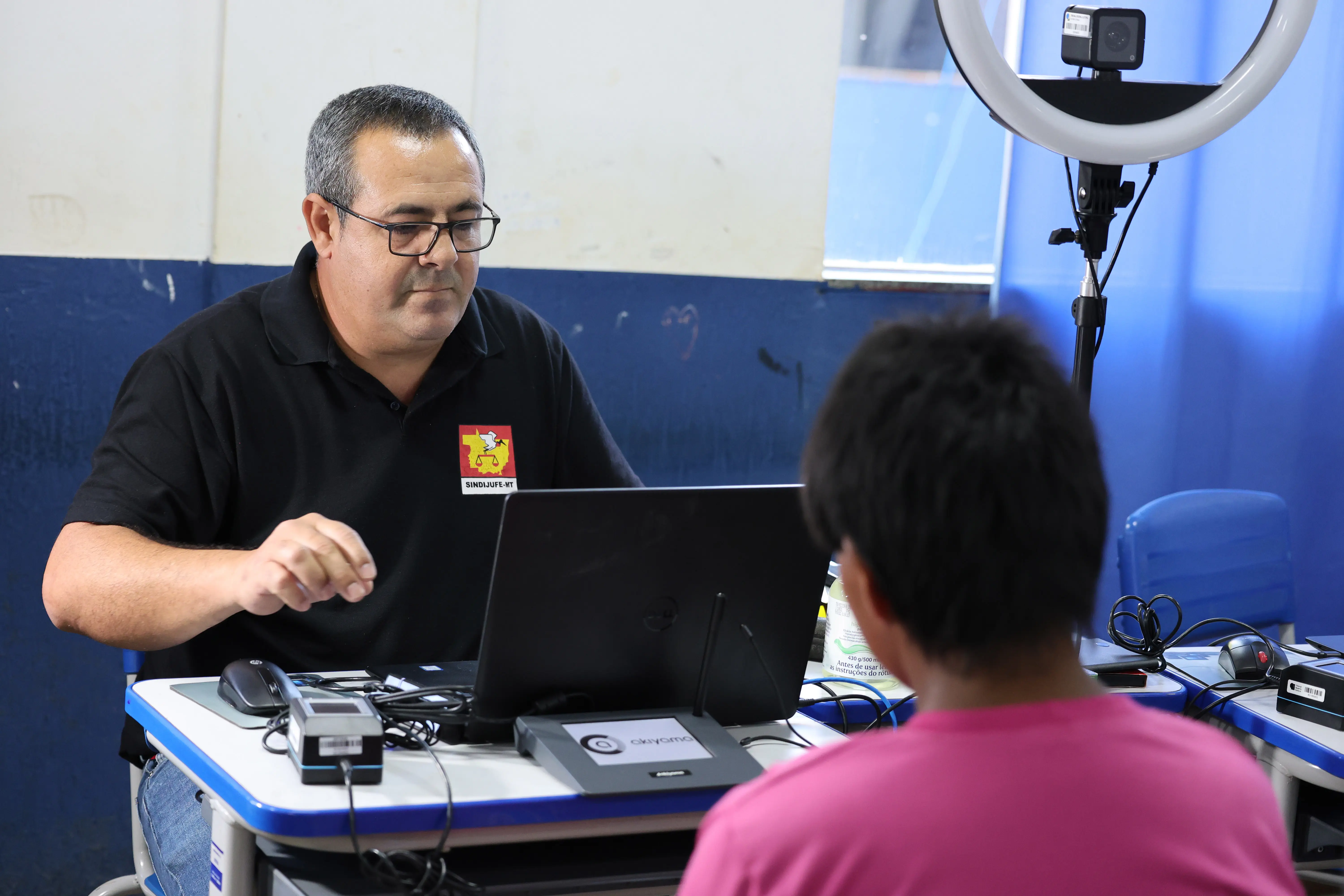 Homem de óculos e camiseta preta com o emblema do Sindijufe-MT atende jovem indígena de blusa rosa em sala azul. Ele utiliza computador e equipamentos de registro biométrico sobre a mesa.