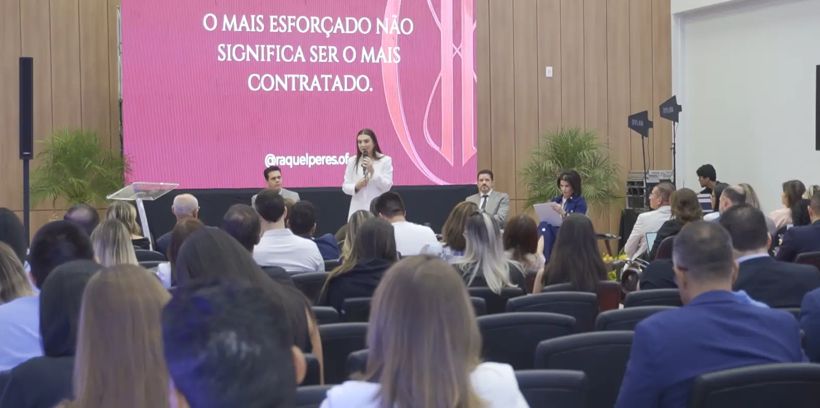 Imagem em plano aberto que mostra várias pessoas de costas, sentadas na plateia. No palco, estão a palestrante Raquel Peres falando ao microfone em pé e, sentados, os mediadores do painel, dentre eles, Jamilson Haddad e Jaqueline Cherulli