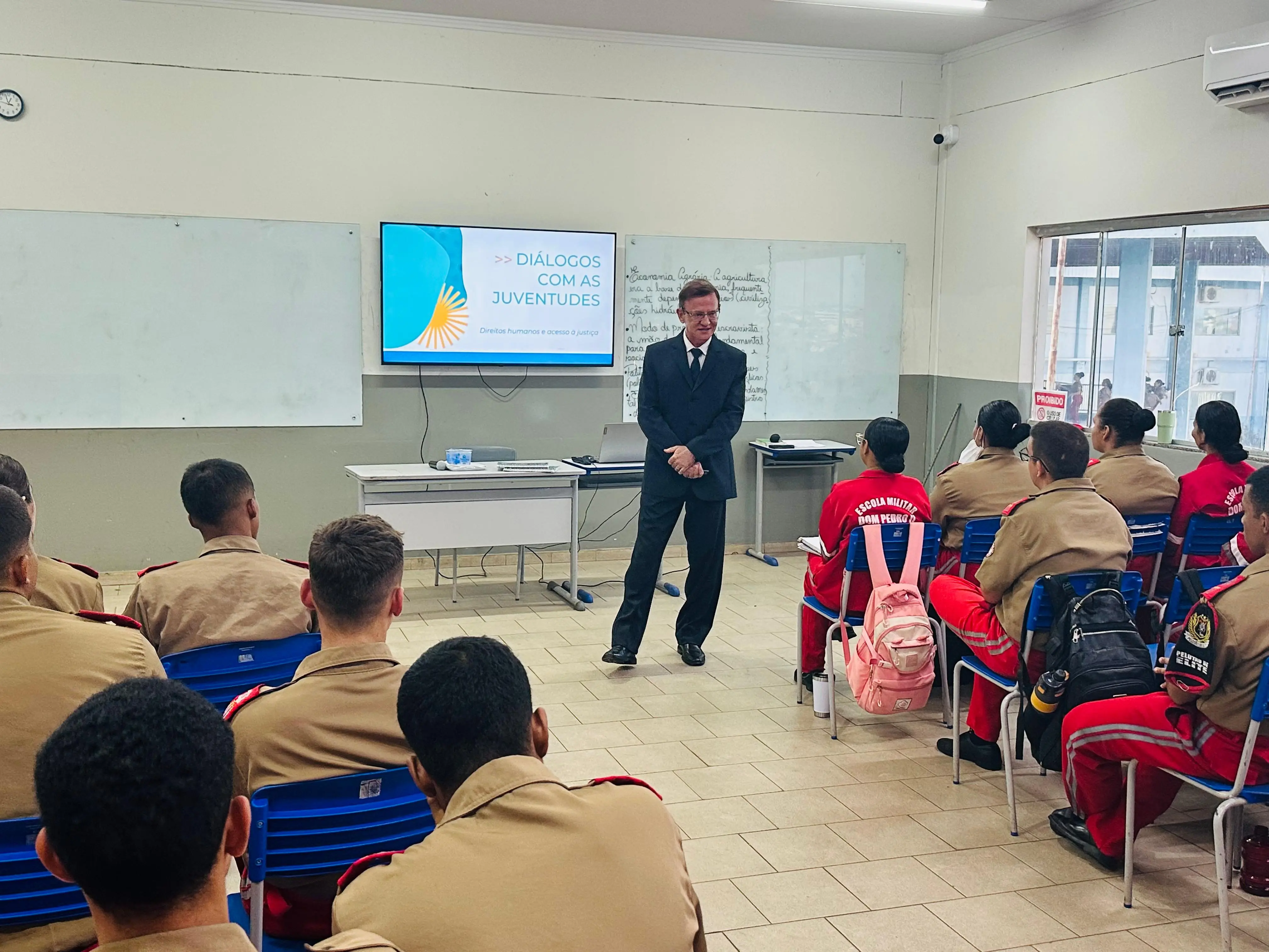 Juiz, de terno preto, é apresentado por bombeiro militar fardado de laranja durante palestra do projeto “Diálogos com as Juventudes”. A atividade ocorre em sala de aula iluminada, com quadro branco e televisão.