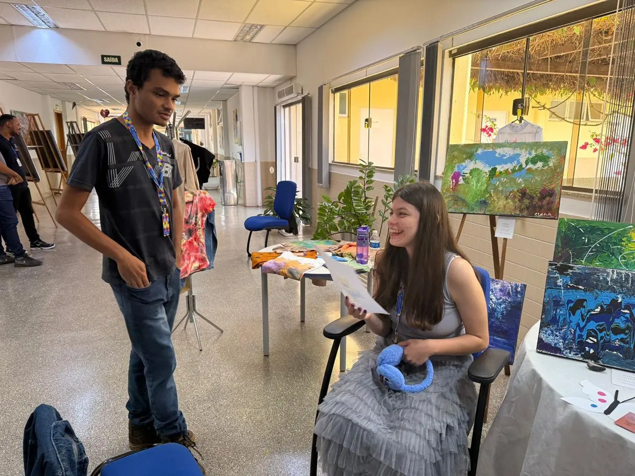 Em um corredor iluminado, Júlio observa com interesse pinturas expostas enquanto conversa com a artista Maria Clara,sorridente. O local é colorido, com obras criativas que destacam talento e expressão artística.