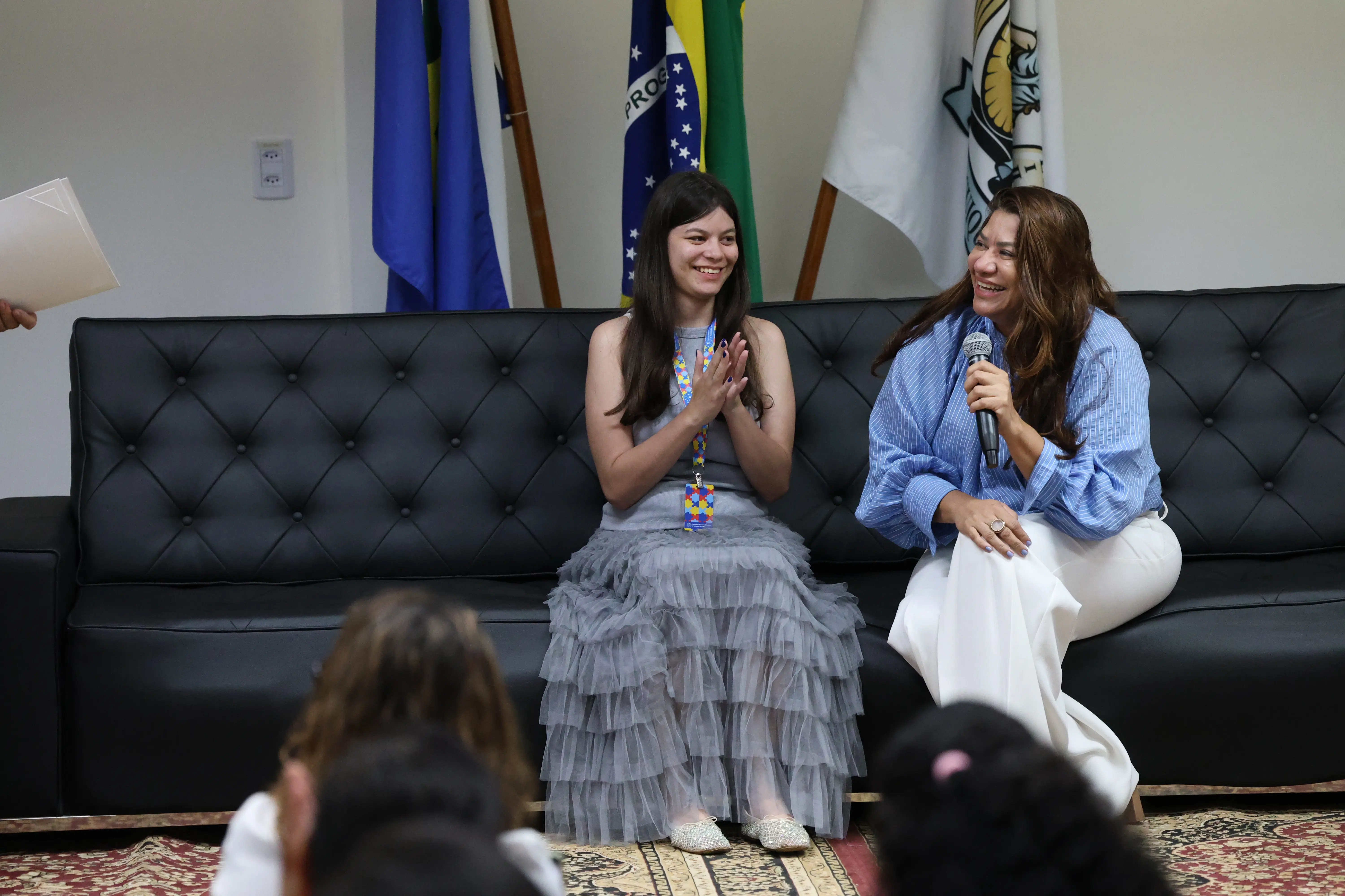 Em auditório, jovem e palestrante sentadas em sofá preto trocam sorrisos e aplausos durante o TJMT Inclusivo. O ambiente é acolhedor, com bandeiras ao fundo e público atento à conversa inspiradora.