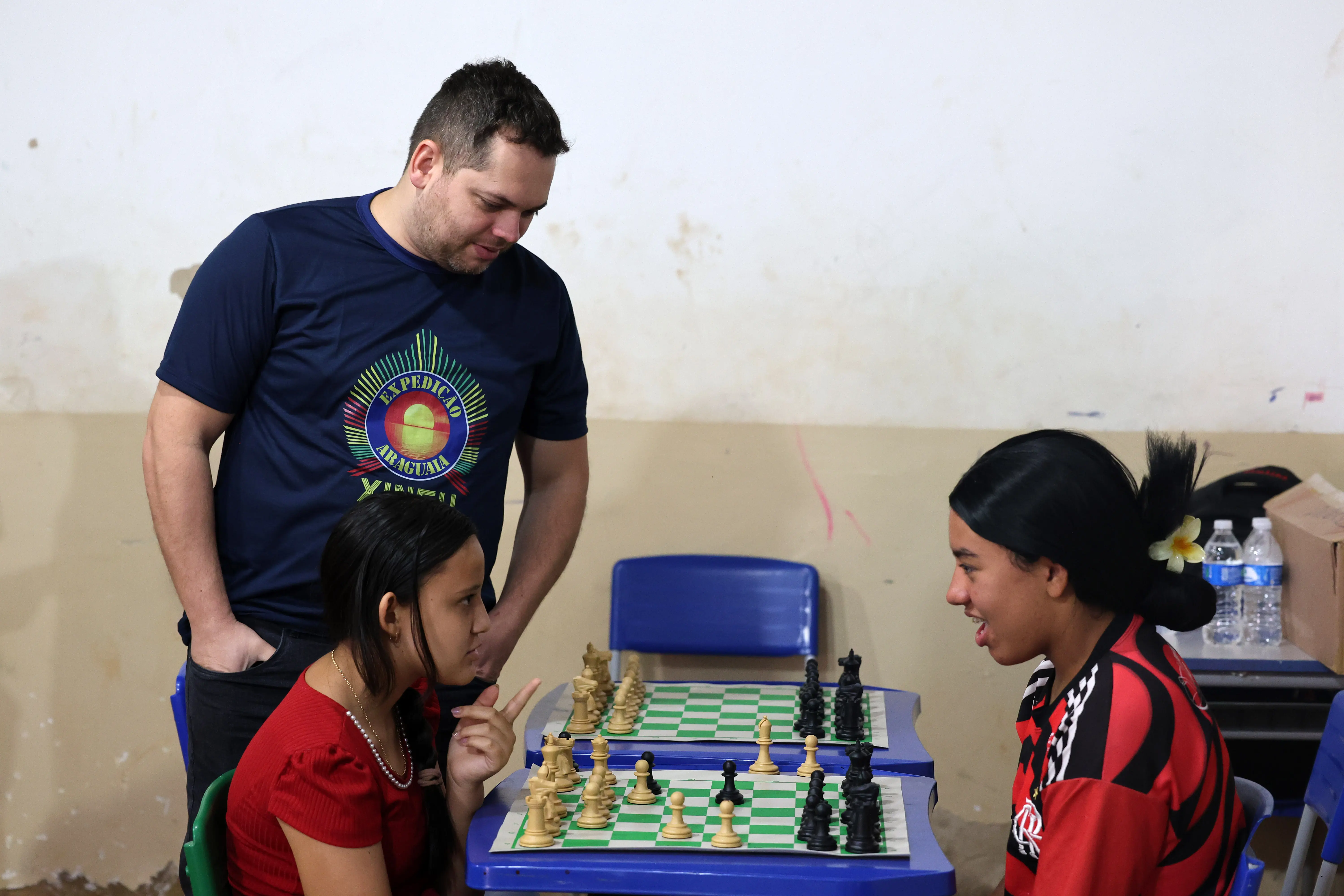 Em sala escolar iluminada, duas jovens jogam xadrez enquanto um servidor da Expedição Araguaia Xingu observa com atenção. O ambiente tem mesas azuis e parede clara, promovendo aprendizado e integração social.