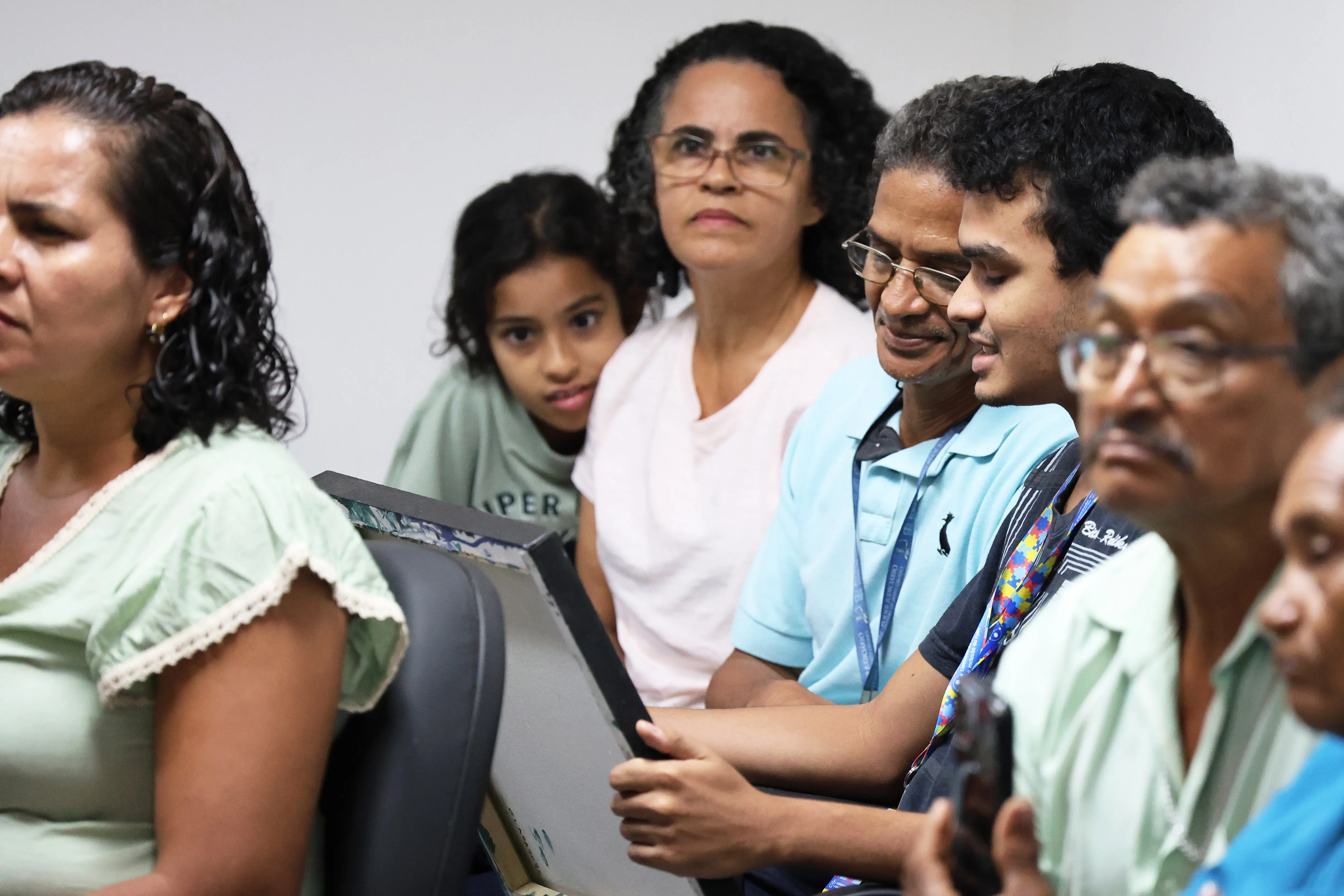 Grupo de pessoas assiste atentamente a uma atividade no TJMT Inclusivo, em Cáceres.Júlio segura uma pintura enquanto é observado por familiares e colegas, em ambiente acolhedor e educativo.