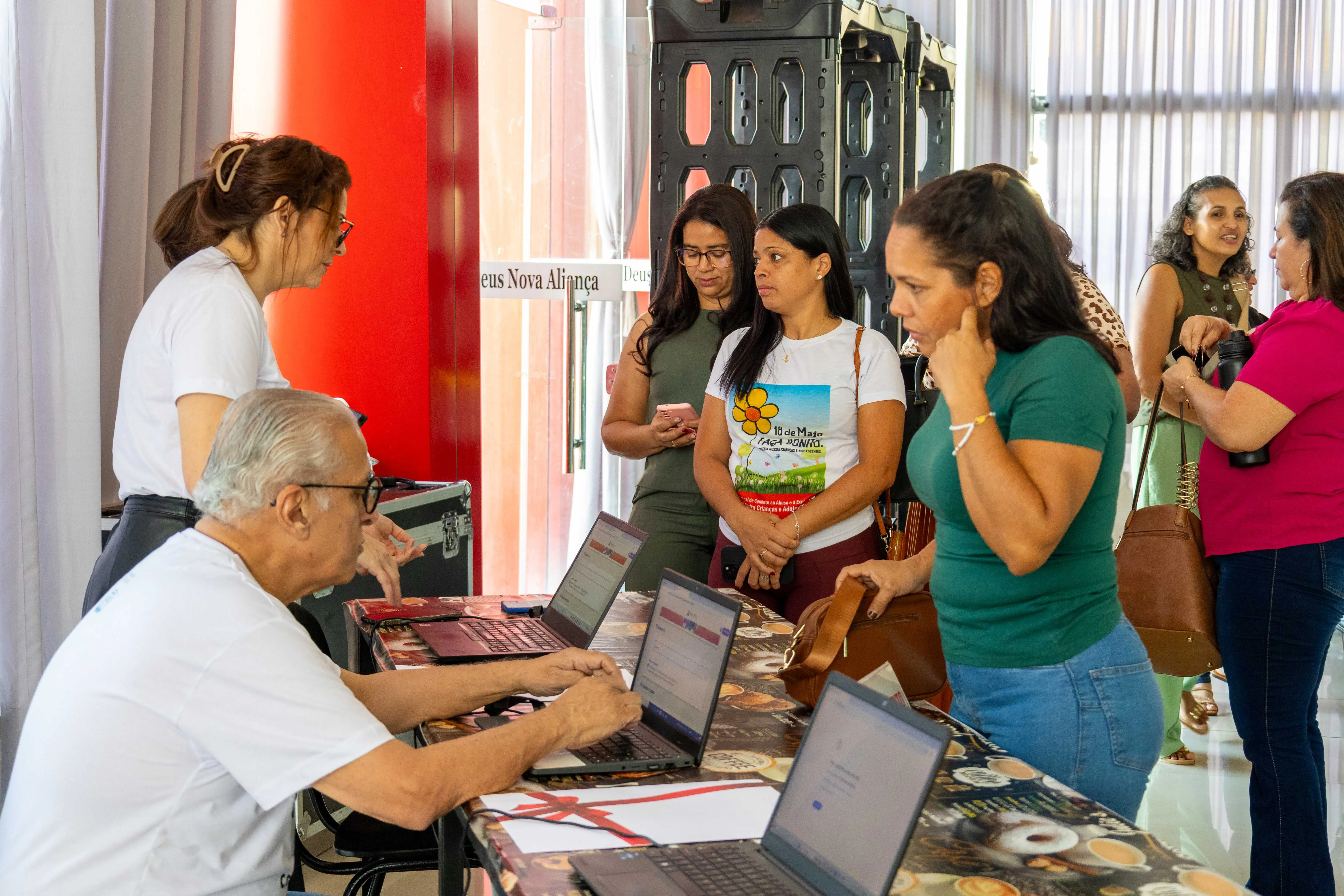 Equipe do evento faz credenciamento de participantes em mesa com notebooks. Homens e mulheres aguardam na fila, conversando em ambiente claro e organizado.