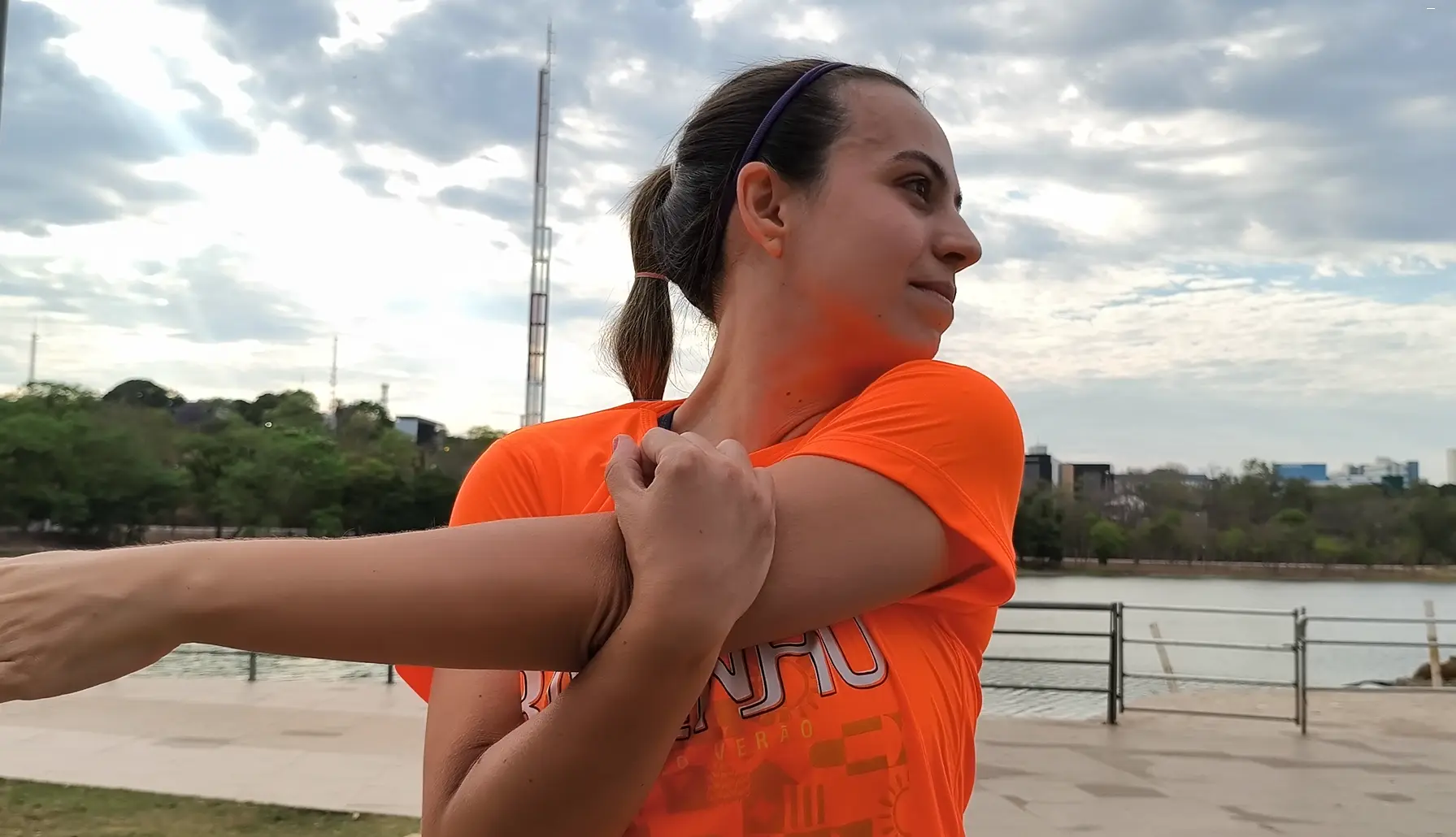 Mulher de camiseta laranja faz alongamento de braço à beira de um lago, com céu nublado e prédios ao fundo.