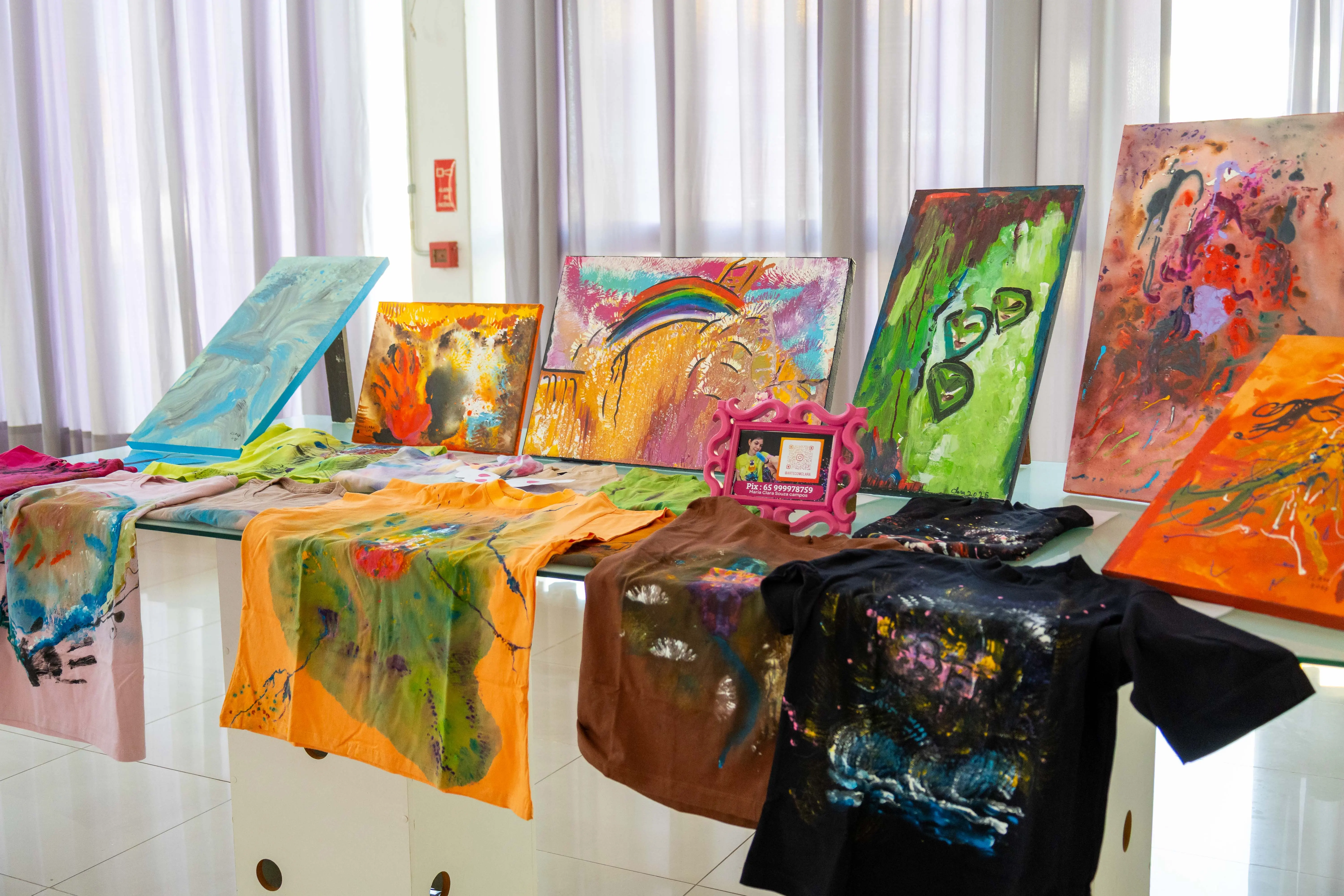 Mesa expositora com camisetas e telas coloridas pintadas à mão, exibindo traços abstratos, arco-íris e texturas vibrantes. O ambiente é iluminado, com cortinas claras ao fundo, remetendo a uma mostra de arte inclusiva e criativa.
