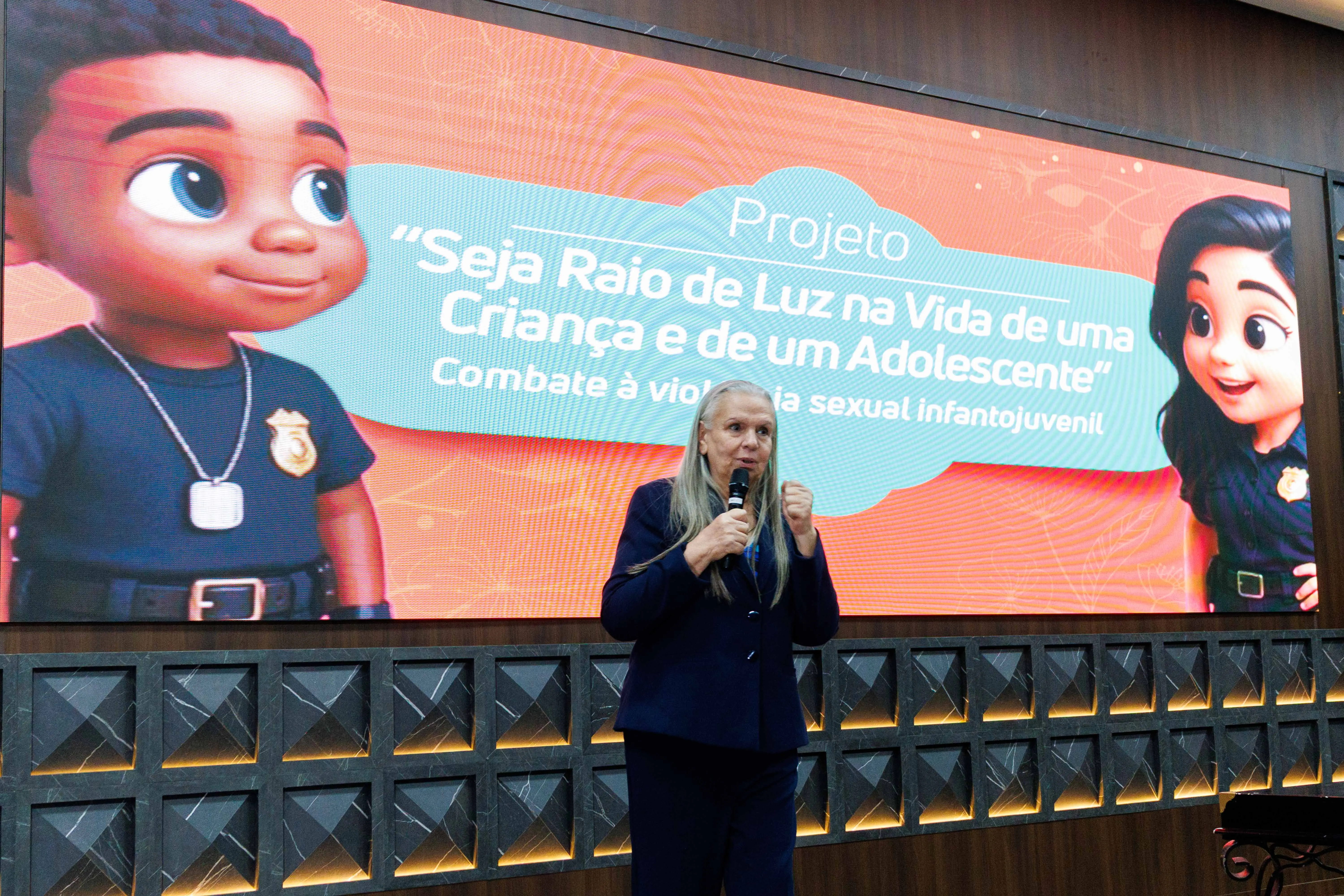 Desembargadora Maria Erotides discursa ao microfone em frente ao painel do projeto “Seja Raio de Luz”. Ilustrações infantis reforçam o tema de proteção. Cena destaca momento de conscientização pública sobre violência sexual.