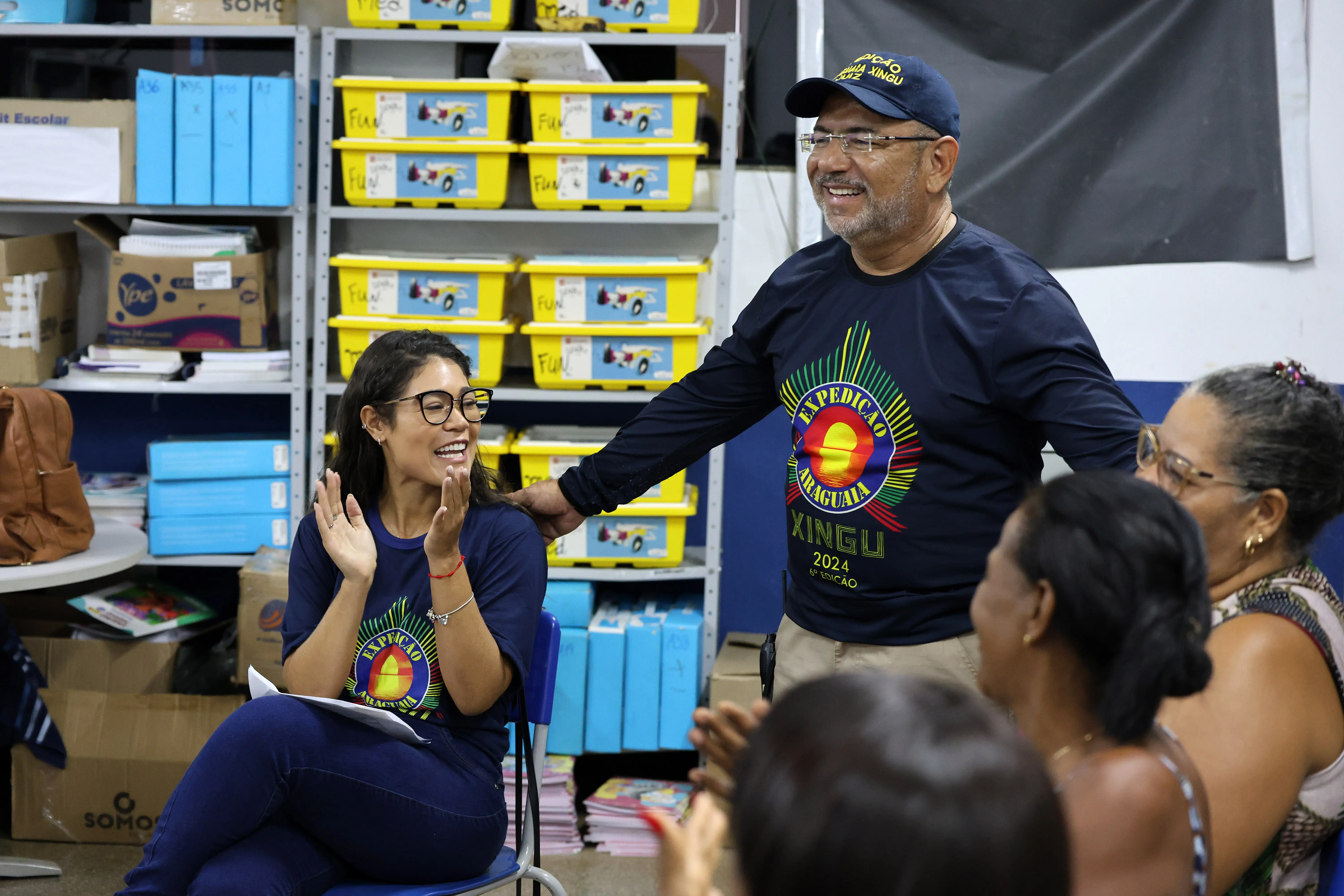 A imagem mostra um grupo reunido em uma sala. Uma pessoa em pé sorri, vestindo camiseta e boné azul com o logo “Experiência Xingu 2024”. Outra, sentada, aplaude. Ao fundo, há prateleiras com caixas amarelas rotuladas “Fichas”.