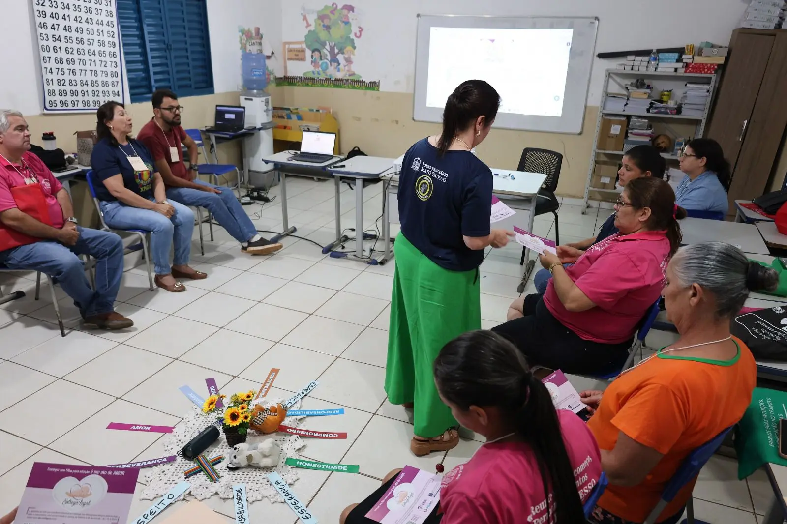Em uma sala simples com piso branco e paredes decoradas com desenhos infantis, pessoas participam de uma roda de conversa sobre valores humanos. No centro, flores e palavras coloridas como “respeito” e “solidariedade” reforçam o clima de acolhimento e reflexão.