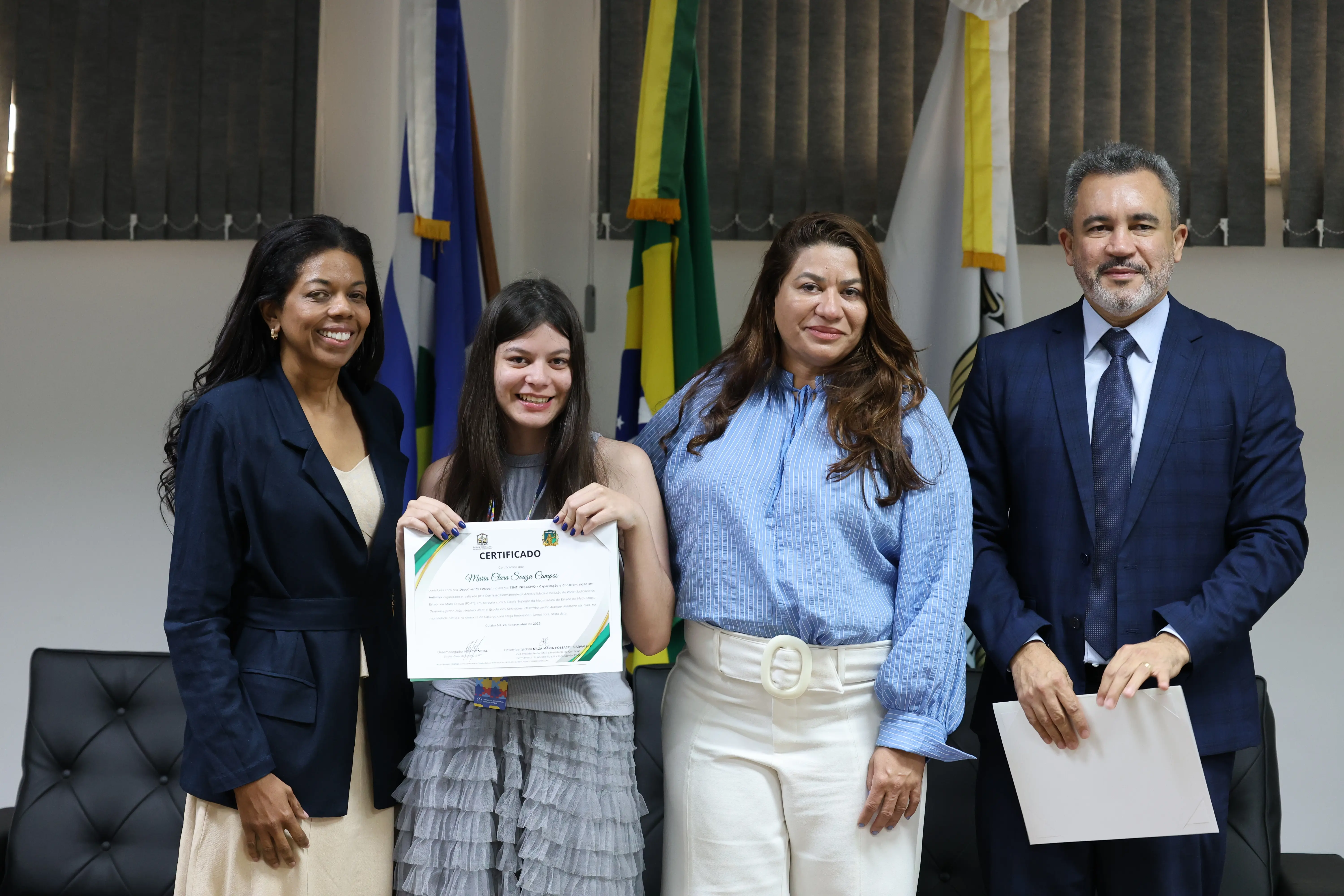 Jovem artista recebe certificado das mãos de representantes do TJMT em cerimônia oficial. As bandeiras ao fundo e os trajes formais reforçam o tom de reconhecimento e valorização da inclusão.