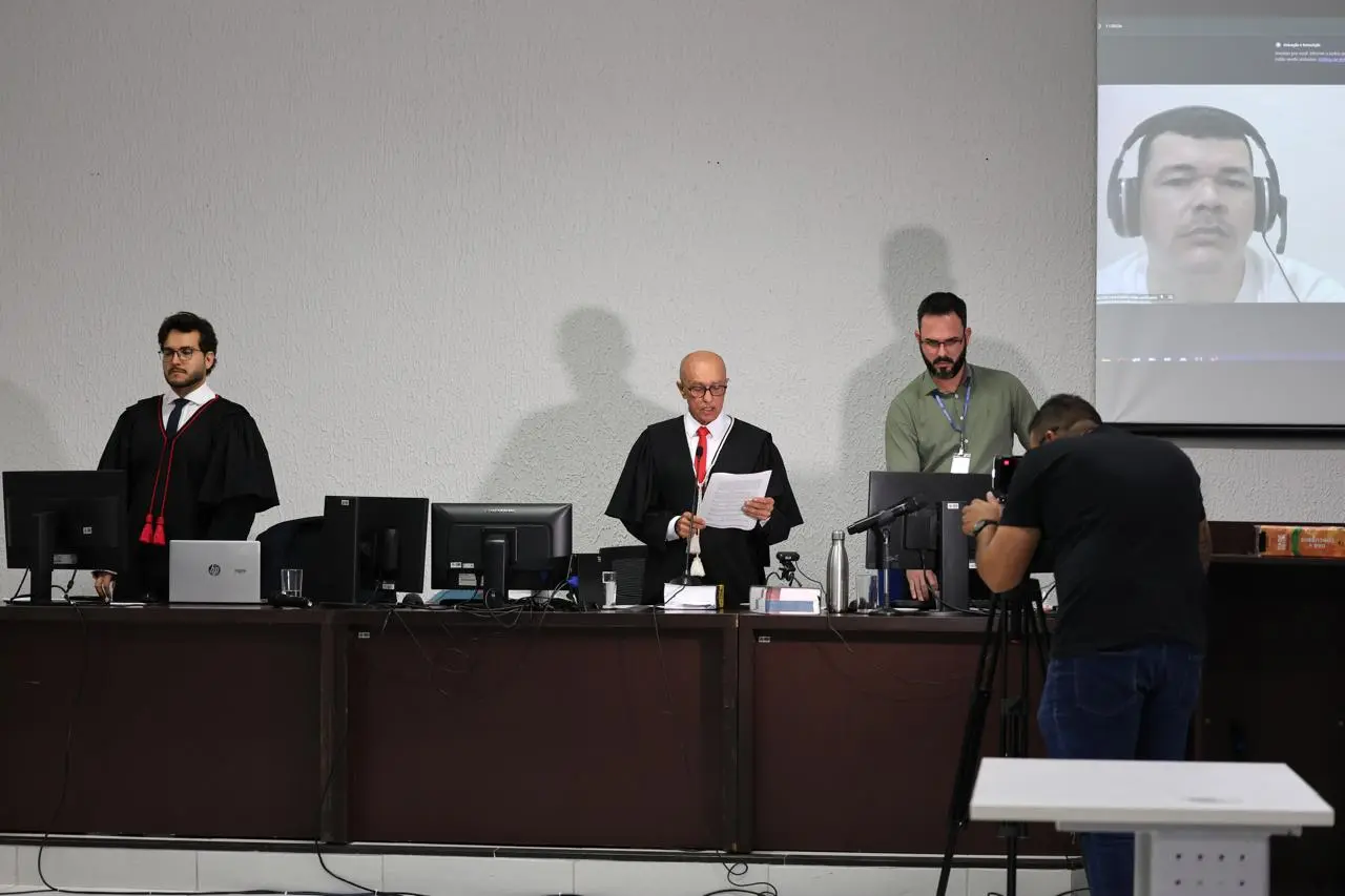 Magistrado realiza a leitura da sentença no plenário do Tribunal do Júri de Sinop, posicionado atrás da bancada, com computadores e microfones. Ao fundo, equipe técnica acompanha a sessão presencial e remota.