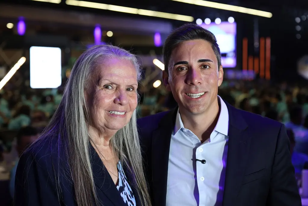 Foto horizontal em plano fechado que mostra a desembargadora Maria Erotides Kneip e o secretário de Educação de Mato Grosso, Alan Porto, posando sorrindo para a foto. Ambos usam blazer azul marinho. Atrás deles, a imagem desfocada é de um grande auditório lotado. 