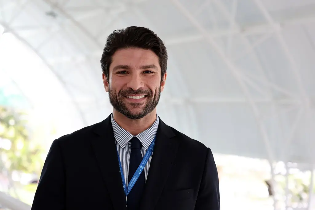 Foto horizontal em plano fechado que mostra o juiz Antônio Bertalia do busto pra cima, sorrindo para a foto. Ele é um homem branco, de cabelos, olhos e barba castanho escuro, usando camisa listrada azul e branca, terno preto e gravata azul marinho.