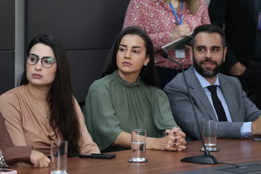 Foto horizontal que mostra duas juízas e um juiz sentados lado a lado, prestando atenção em uma apresentação. No centro está a juíza Gabriela Andressa Moreira Dias. Ela é uma mulher jovem, parda, de cabelos longos e castanhos, usando blusa de gola alta na cor verde militar.