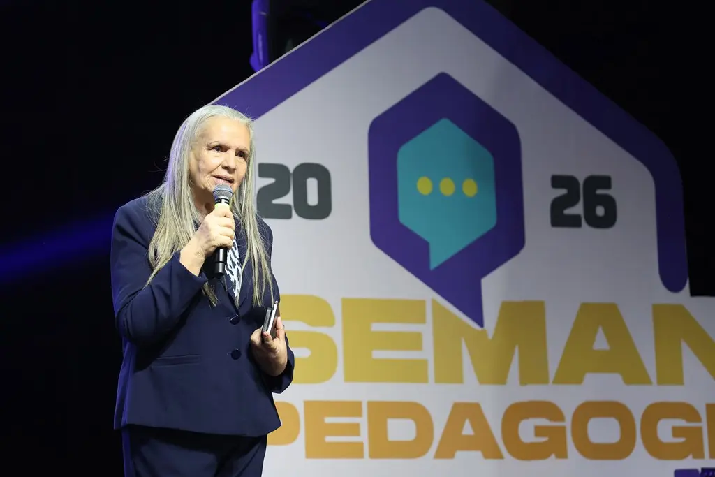 Foto horizontal em plano médio que mostra a desembargadora Maria Erotides Kneip em pé, falando ao microfone, no palco da Semana Pedagógica 2026. Ela é uma senhora branca, de cabelos longos e grisalhos, usando calça e blazer azul marinho. Atrás dela, aparece a logomarca da Semana Pedagógica 2026. 