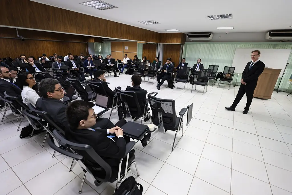 Foto horizontal em plano aberto que mostra uma sala de aula com juízes e juízas sentados em carteiras dispostas em formato de U. O projetor, juiz Wanderlei Reis, está à frente, em pé. 