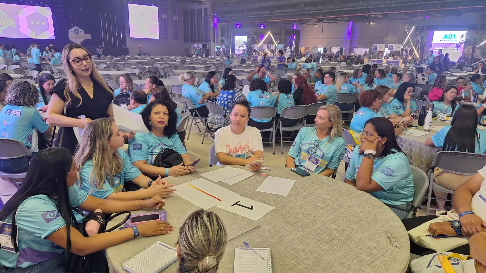 Foto horizontal ambientada em um grande salão de eventos. Em primeiro plano, professores usando camiseta da Semana Pedagógica estão sentados em volta de uma mesa redonda. Em pé, a juíza Ana Graziela Vaz orienta sobre a oficina sobre violência doméstica. 