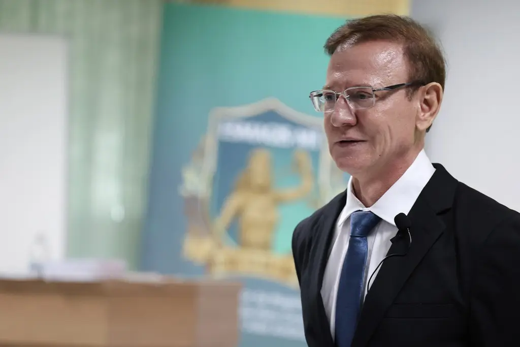  Foto horizontal em plano fechado que mostra o juiz Wanderlei Reis de busto pra cima, de perfil, durante sua aula. Ele é um homem branco, de cabelos e olhos castanhos claros, usando camisa branca, terno preto, gravata azul, óculos de grau e microfone de lapela. 