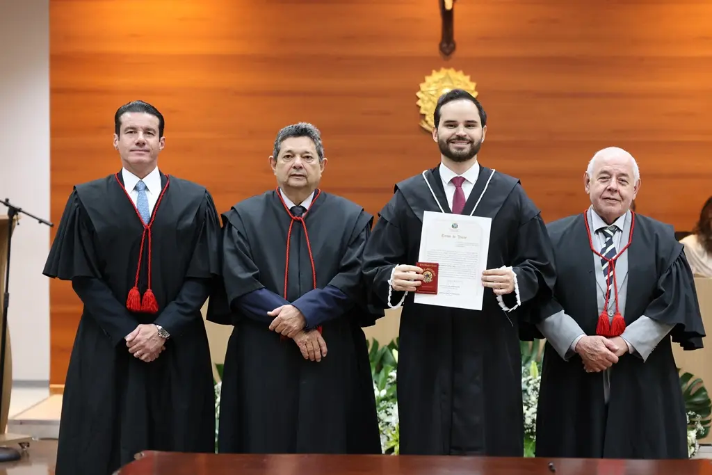 Foto do juiz Israel Tibes ao lado do presidente, do corregedor-geral e do ouvidor do TJMT, durante sessão de posse. Israel segura seu termo de posse e sua carteira funcional. Todos usam toga e sorriem para a foto.