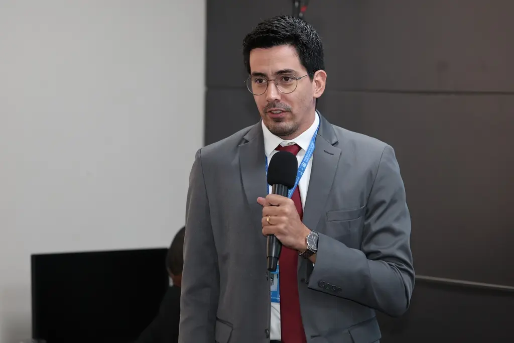 foto horizontal que mostra o assessor de Assuntos Previdenciários do TJMT, Bruno Santana, em pé, falando ao microfone. Ele é um homem branco, de olhos e cabelos escuros, usando camisa branca, gravata vermelha, terno cinza, crachá funcional e óculos de grau.