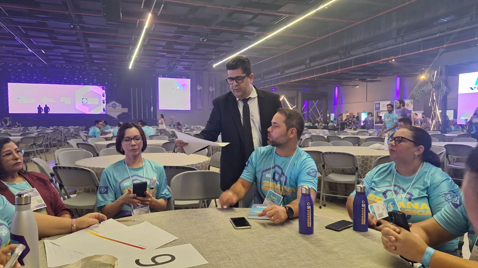 Foto horizontal ambientada em um grande salão de eventos. Em primeiro plano, professores usando a camiseta da Semana Pedagógica estão sentados em volta de uma mesa redonda. Em pé, o juiz Marcos Terêncio os orienta sobre a oficina sobre violência doméstica.