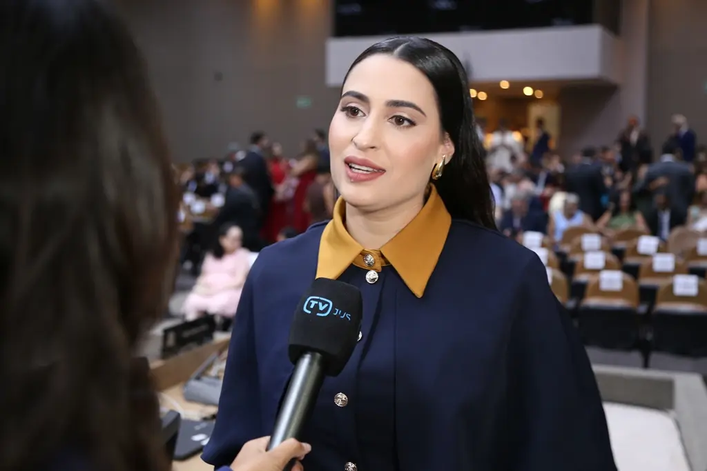 Foto horizontal da juíza Ana Carolina Pelicioni da Silva Volkers durante entrevista à TV Justiça. ela é uma mulher branca, de cabelos pretos e lisos, usando vestido azul marinho com gola amarelo queimado.