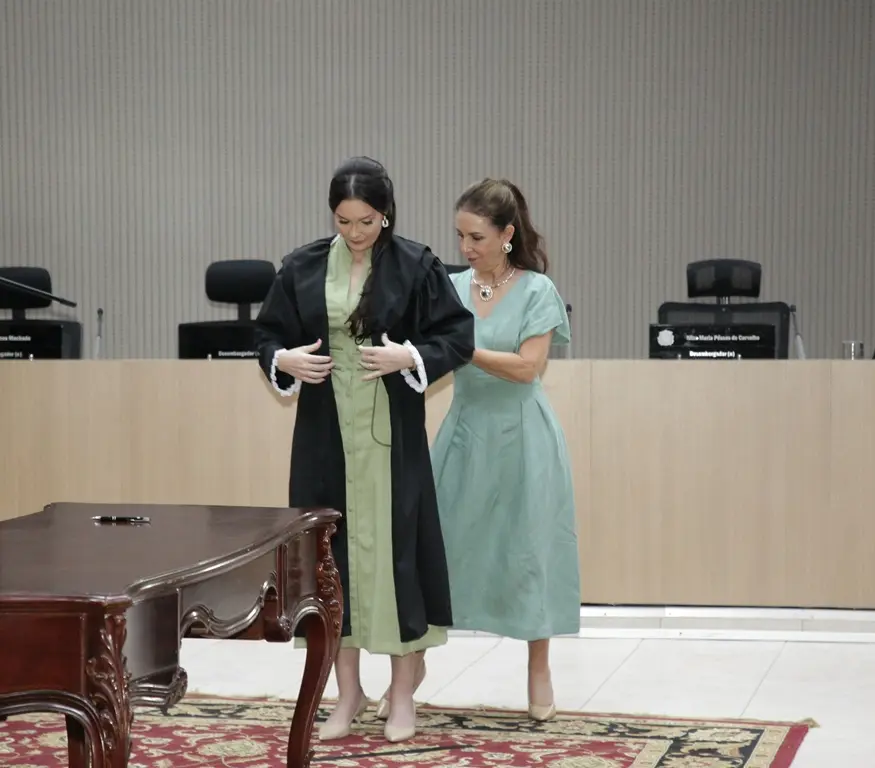 Foto horizontal que mostra a juíza Luana Wendt sendo auxiliada por sua mãe para vestir a toga, durante sessão de posse. Ambas usam vestido verde e estão no centro do Plenário 1 do TJMT.