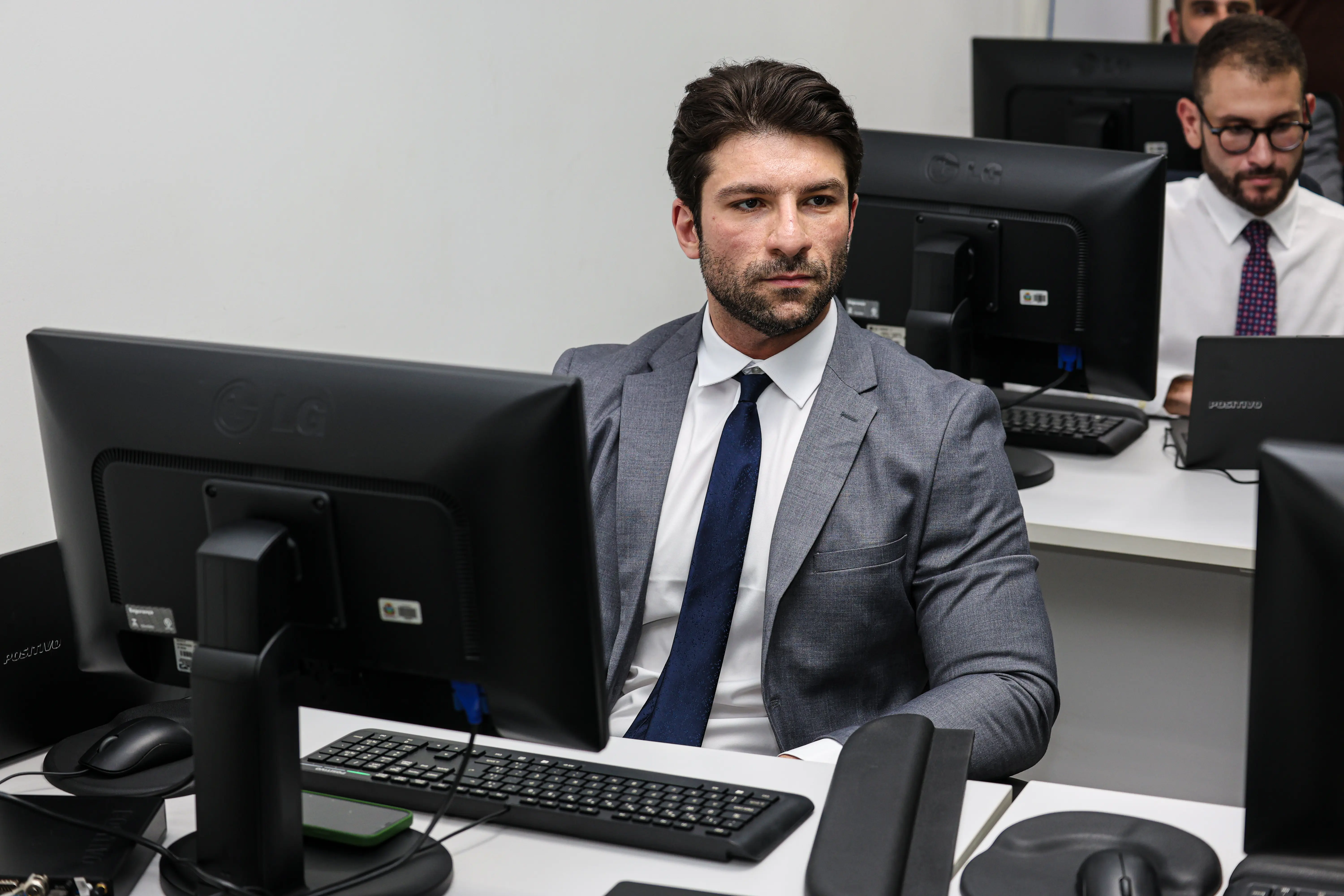 Homem de terno cinza e gravata azul acompanha atividade em laboratório de informática, atento à tela do computador, durante capacitação técnica.
