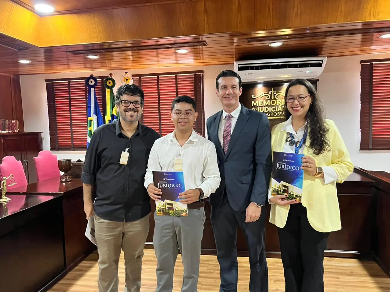 Quatro pessoas posam para foto em sala institucional. Dois deles seguram exemplares do Glossário Jurídico. Ao fundo, placa com os dizeres 