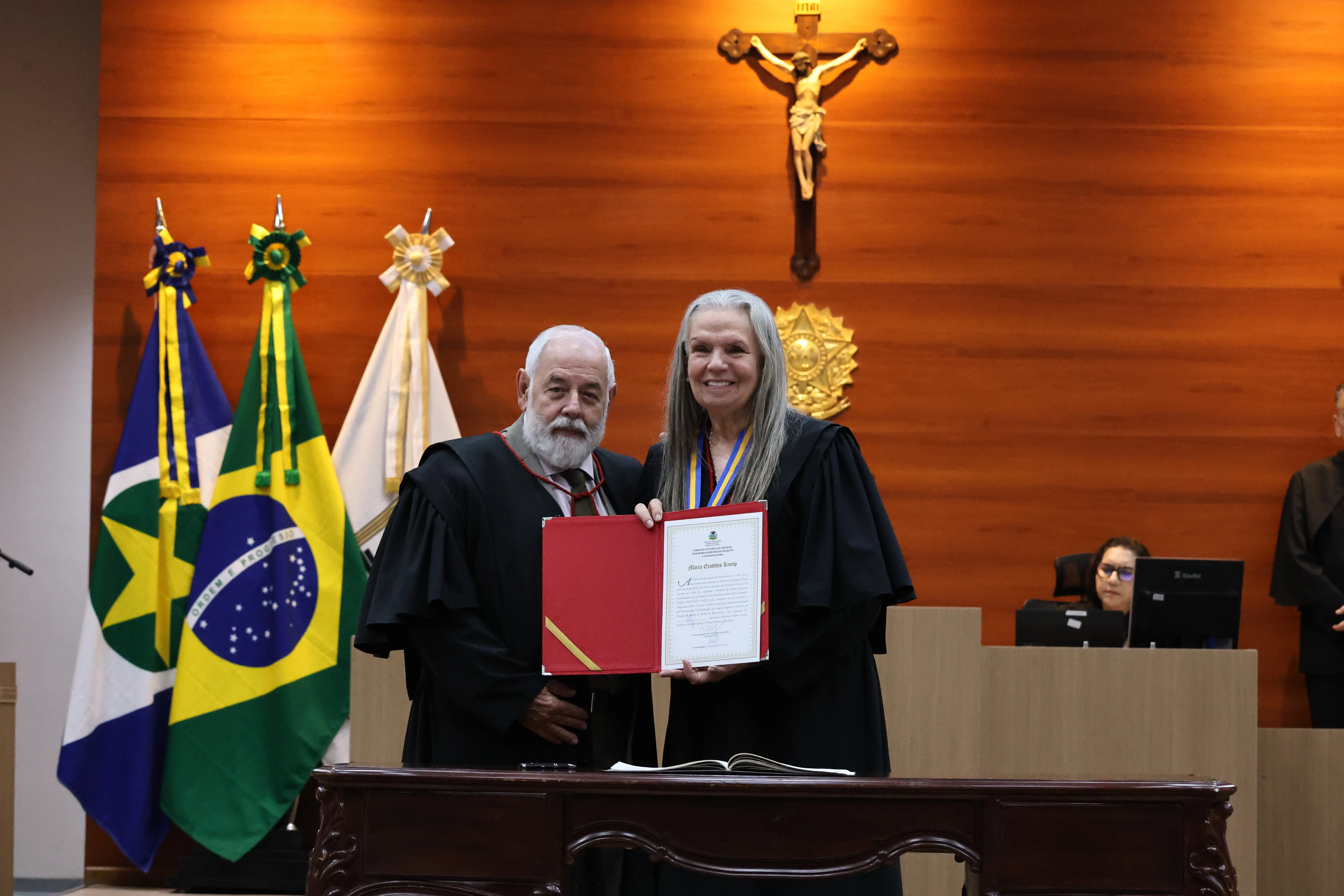 Homem e mulher de toga seguram juntos um diploma em pasta vermelha. Ao fundo, painel de madeira com um crucifixo e as bandeiras de Mato Grosso e do Brasil. Uma pessoa sentada ao computador.