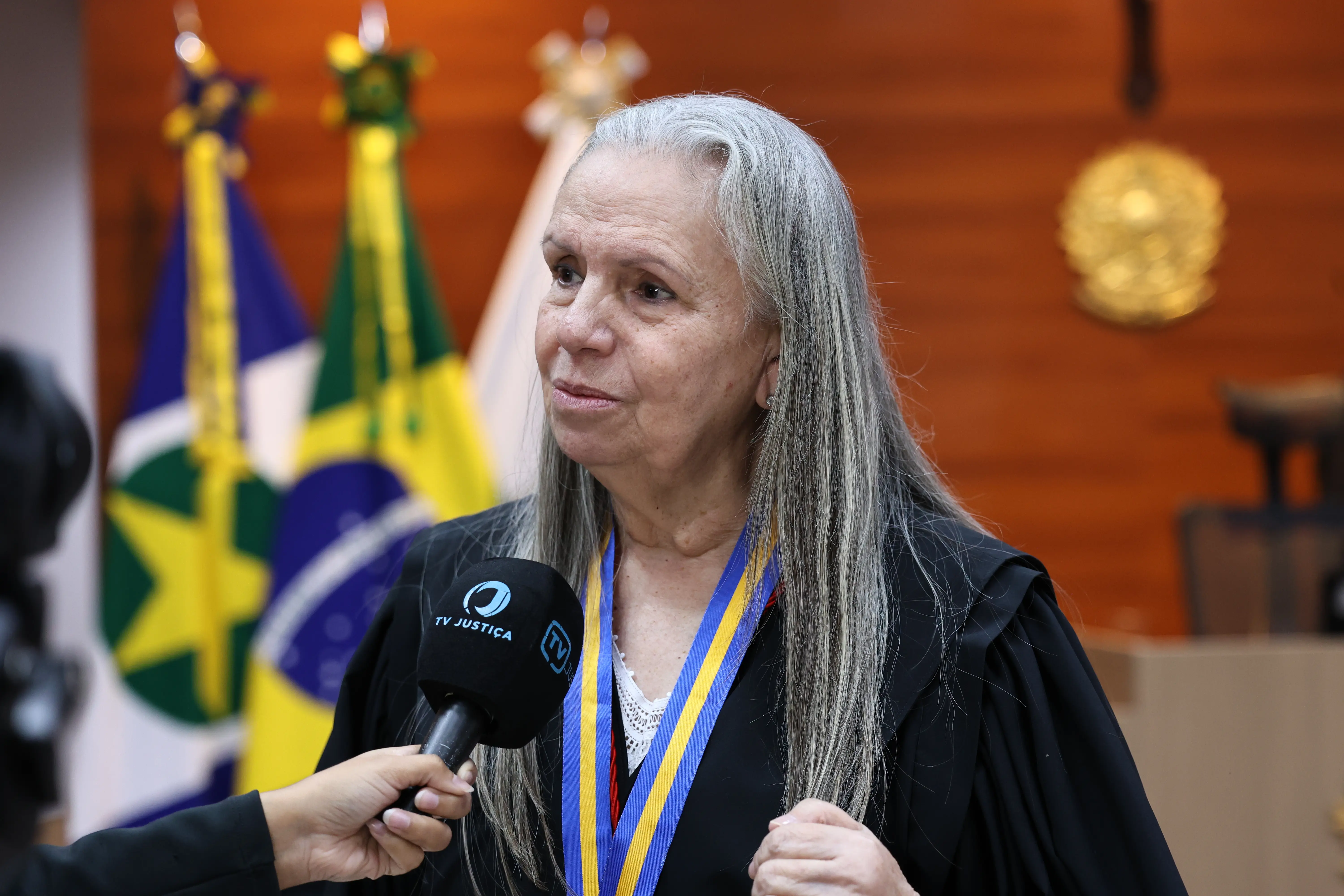 Mulher branca de longos cabelos grisalhos e toga preta concede entrevista ao microfone da 