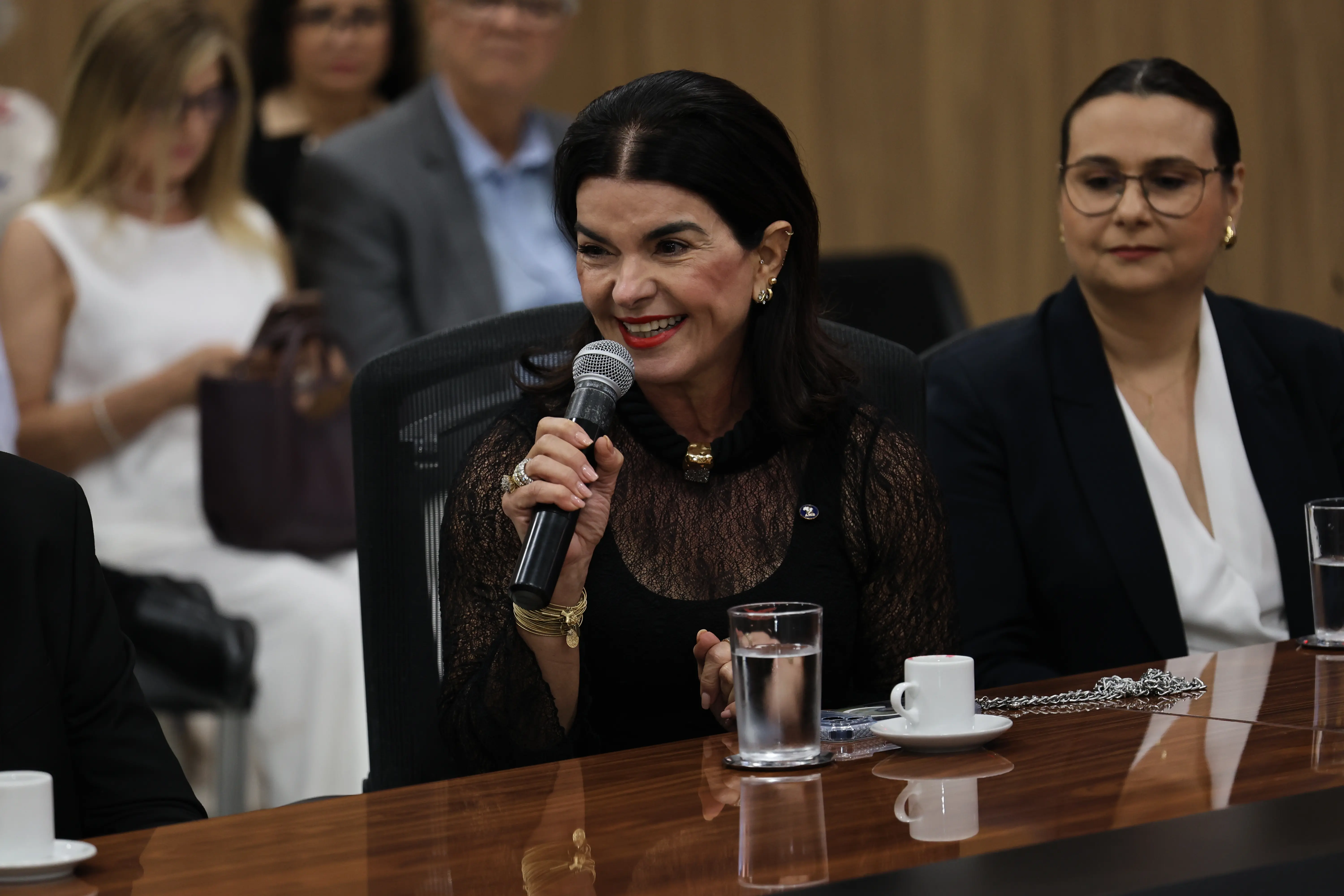 Mulher de cabelos escuros e blusa preta rendada sorri ao falar num microfone. À direita, outra mulher de óculos e blazer preto observa. Na mesa de madeira há copos d'água e uma xícara de café.
