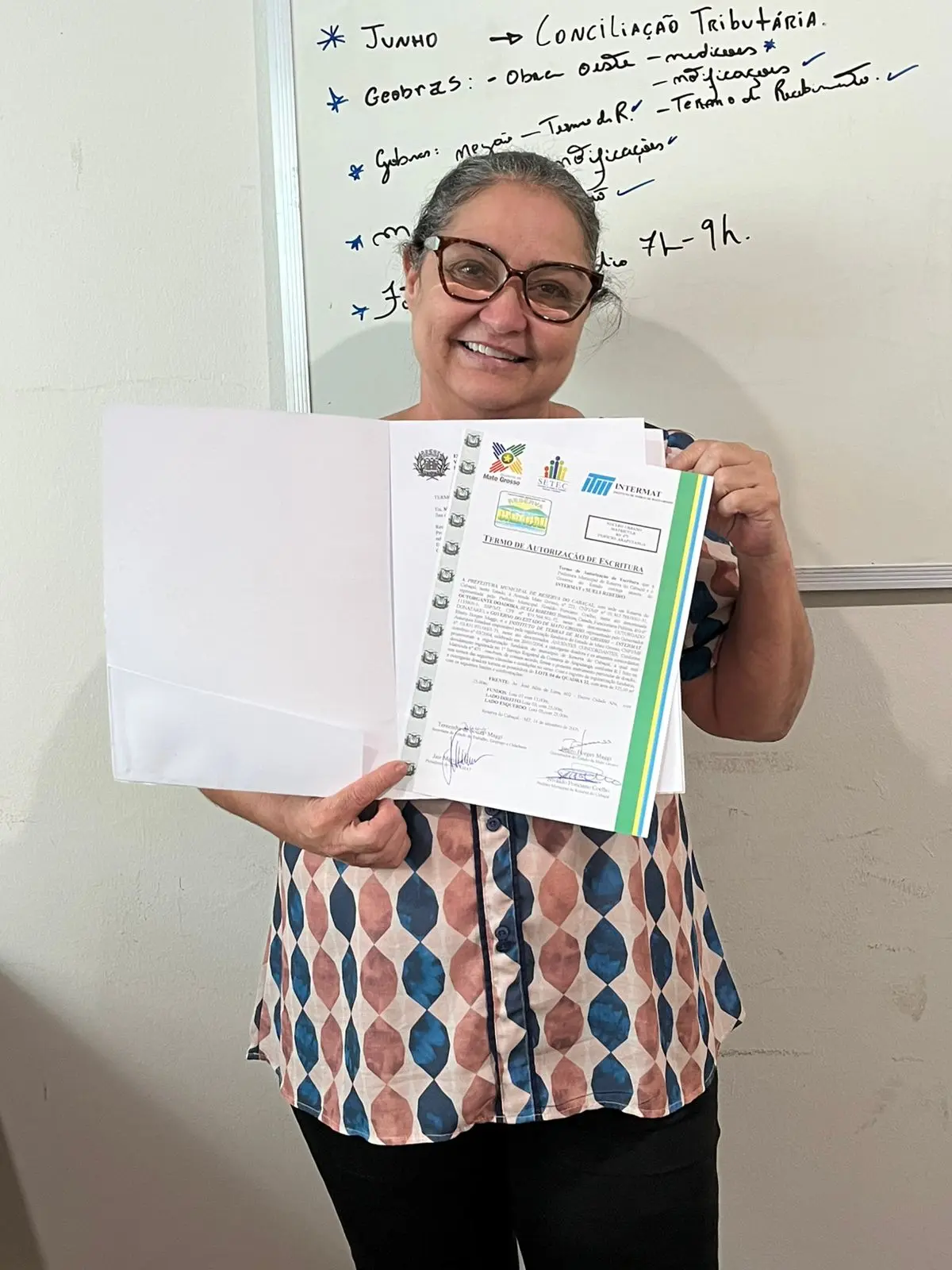 Foto vertical que mostra uma senhora mostrando o título de propriedade de imóvel, sorrindo. Ela usa óculos e blusa estampada. 