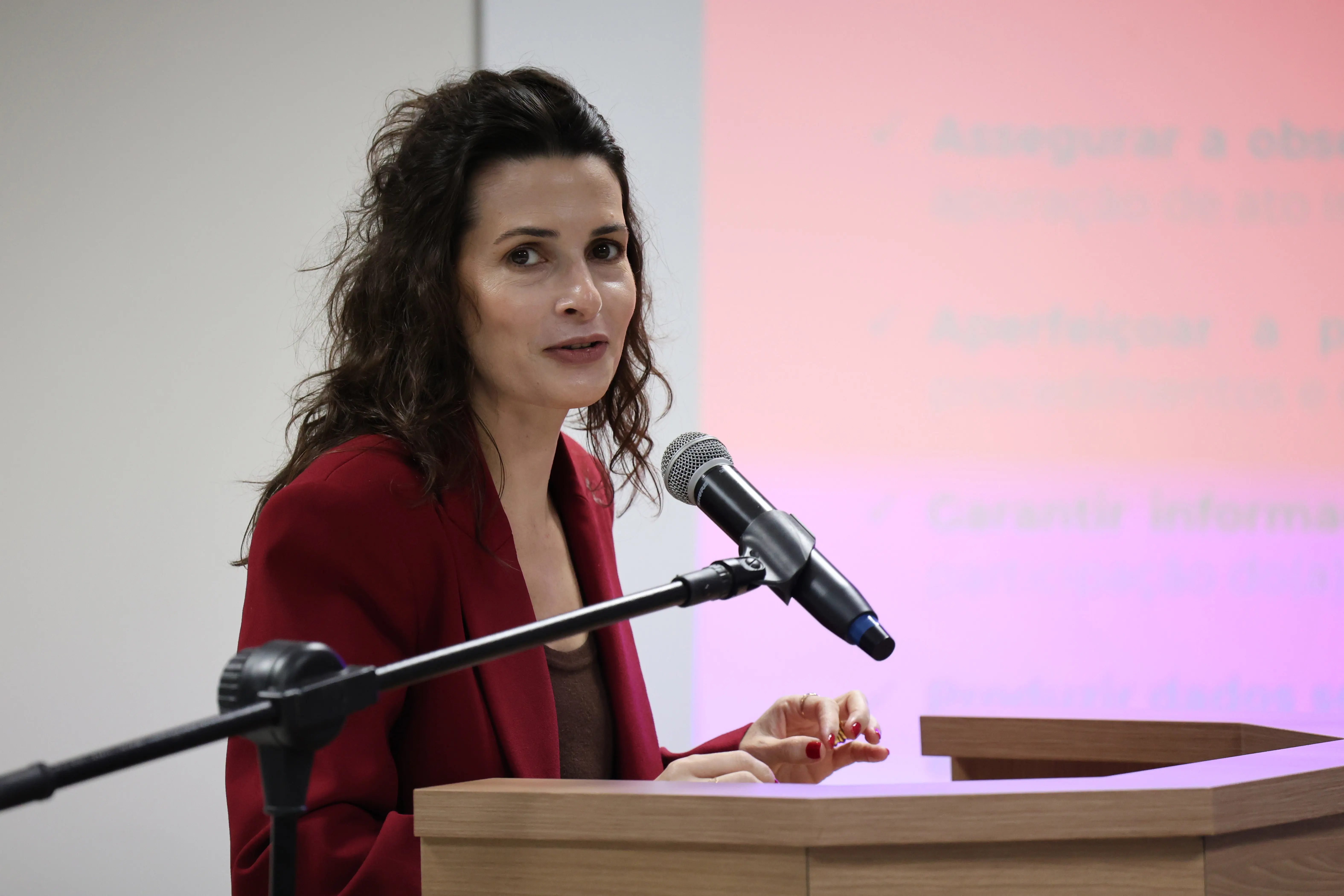Mulher branca de cabelos escuros e terno vermelho fala ao microfone em um púlpito de madeira. Ela olha para frente com expressão serena. Ao fundo, parte de uma tela projeta um texto em rosa claro.