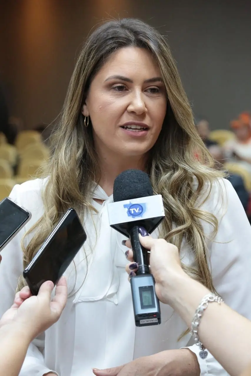Mulher loira de blusa branca concede entrevista. Duas mãos seguram celulares perto dela, e uma terceira mão segura um microfone com a logo 