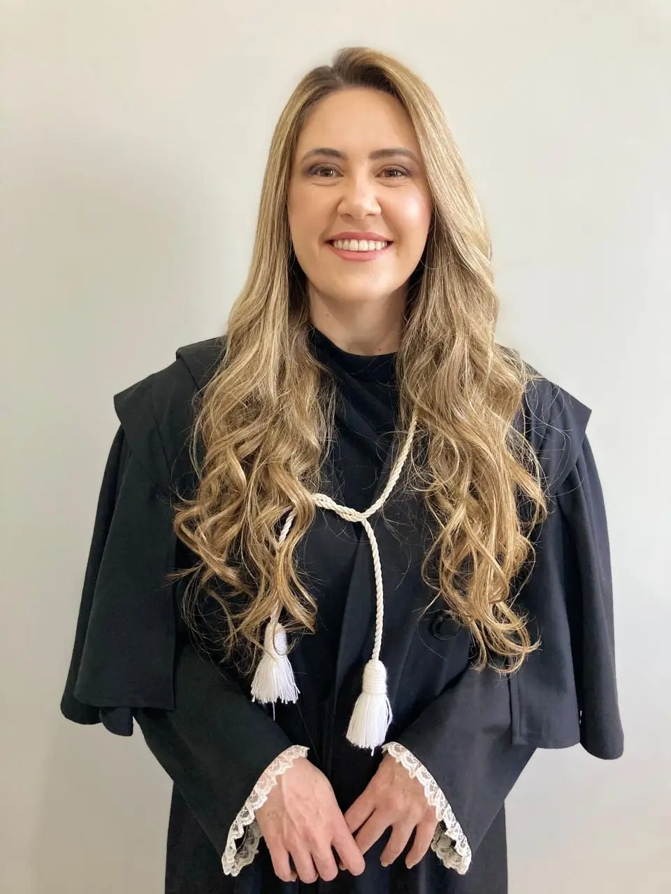 Mulher loira sorri de frente para a foto vestindo uma toga jurídica preta com cordão e borlas brancas. Suas mãos estão cruzadas à frente sobre o fundo branco.
