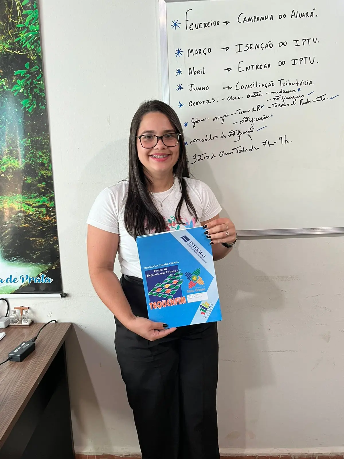 Foto vertical que mostra uma mulher jovem sorrindo e segurando a pasta com o título de propriedade de imóvel. Ela tem a pele clara, cabelos longos, lisos, soltos e castanhos, usando camiseta branca, calça preta e óculos de grau.