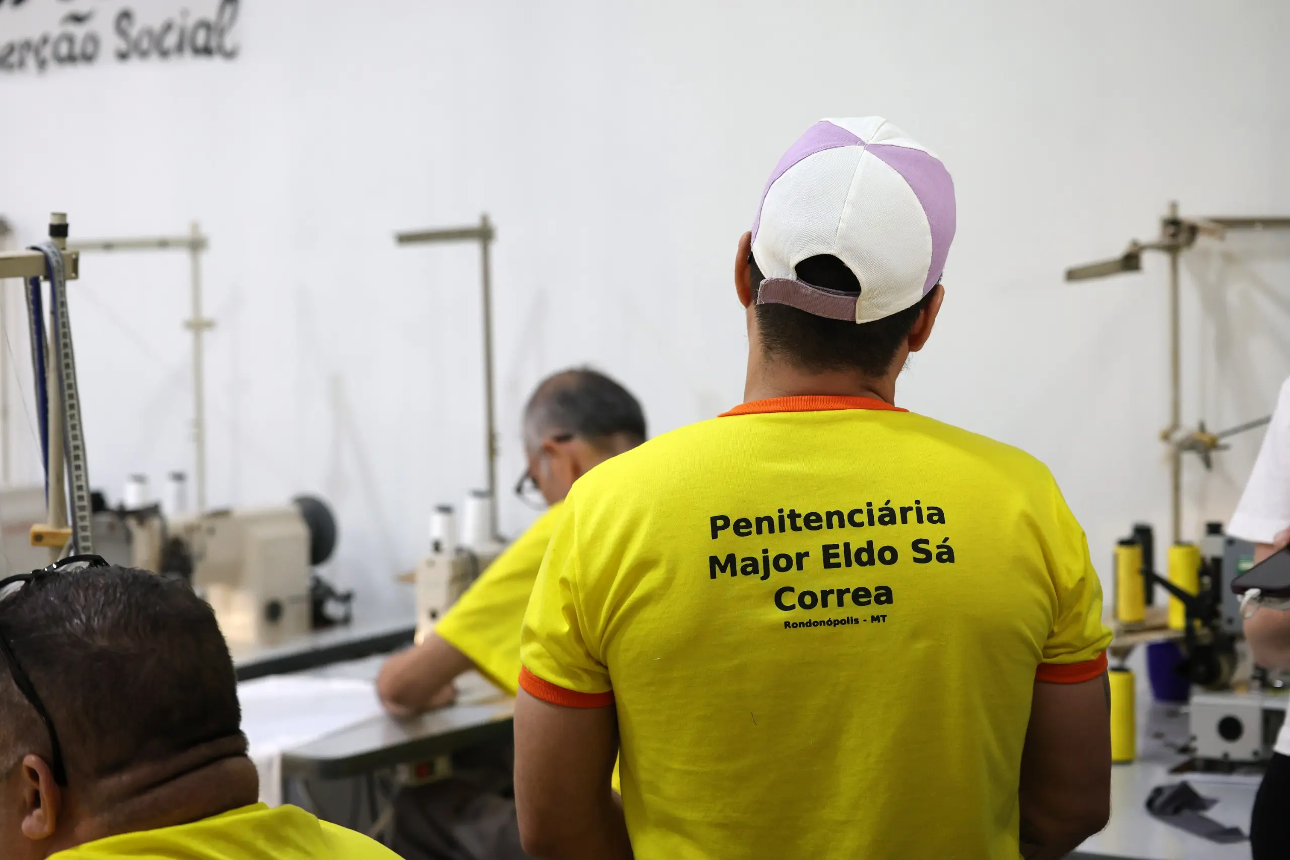 Homem de costas veste camiseta amarela com a inscrição "Penitenciária Major Eldo Sá Correa - Rondonópolis - MT" e boné branco e lilás. Ele está em uma sala com máquinas de costura.