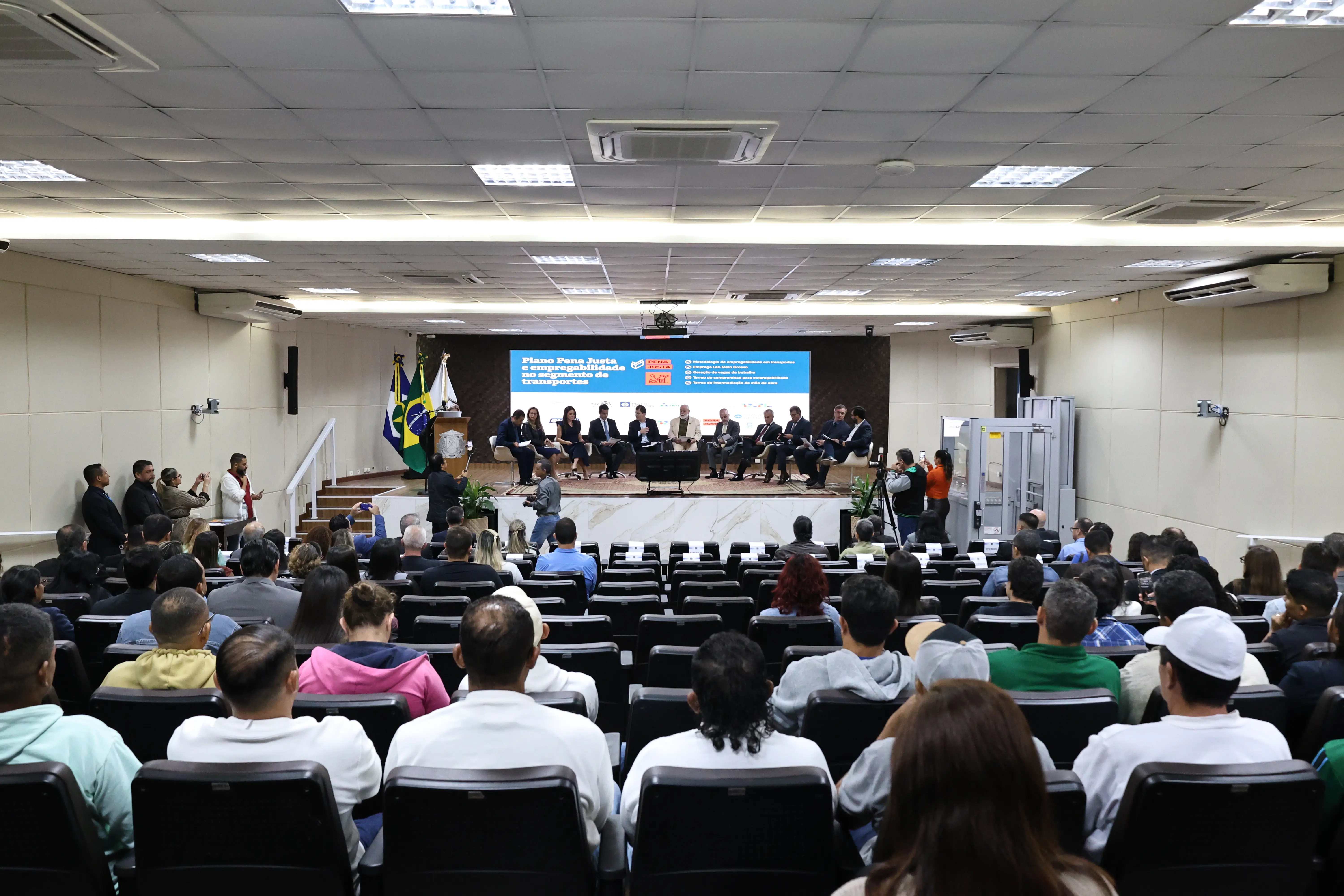 Auditório com iluminação clara, com dezenas de pessoas. Algumas sentadas em poltronas, outras no palco, em frente a um telão em azul com letras brancas. 