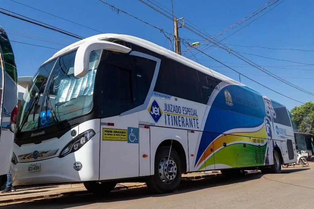 Ônibus branco estacionado sob céu azul. Na lateral, as inscrições "Juizado Especial Itinerante" e logos do Poder Judiciário de Mato Grosso em tons de azul, verde e amarelo.