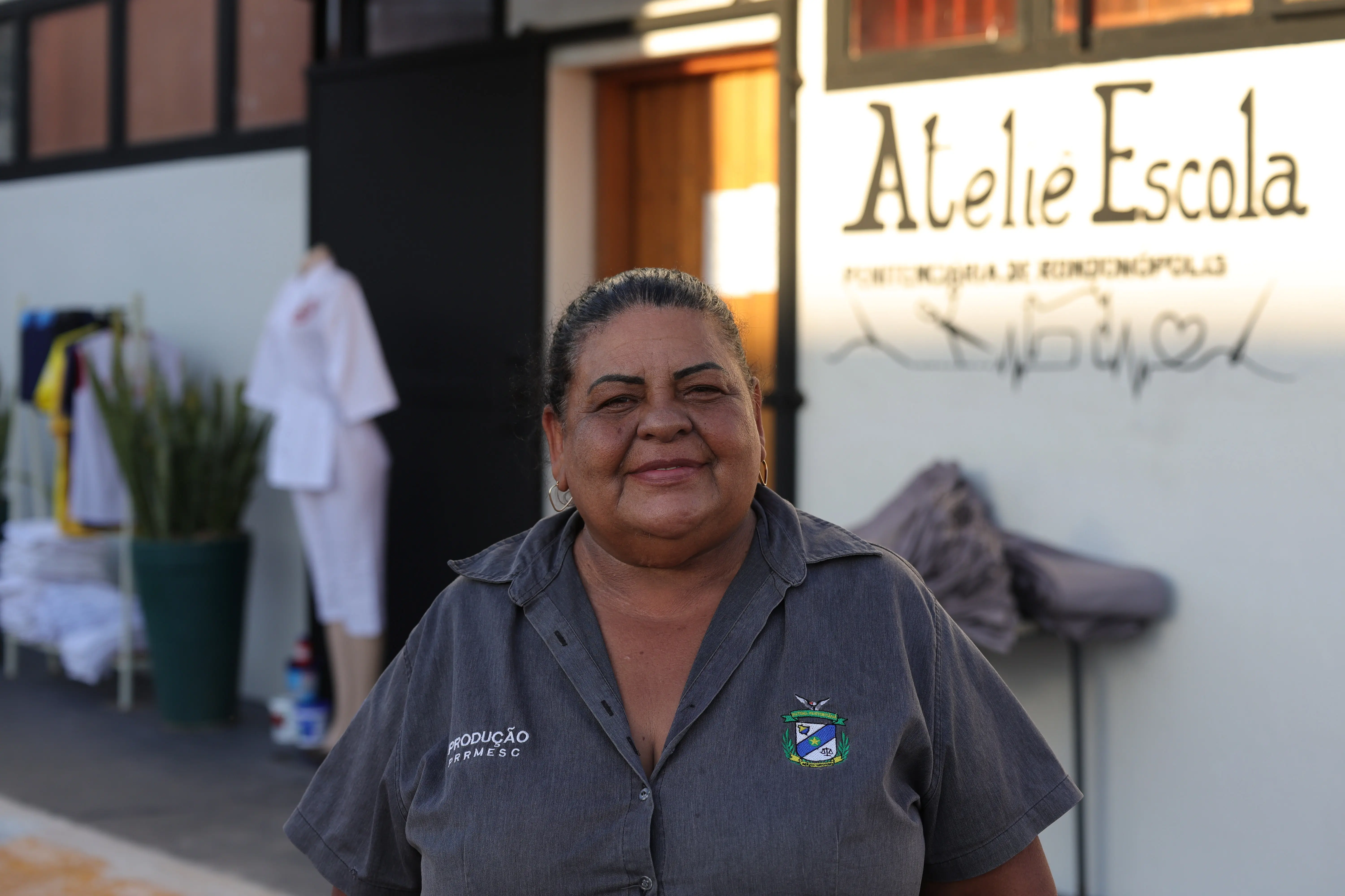 Retrato de uma mulher de cabelos presos, sorrindo em frente a uma parede branca com a inscrição "Ateliê Escola". Ela veste uma camisa cinza com um brasão estampado.