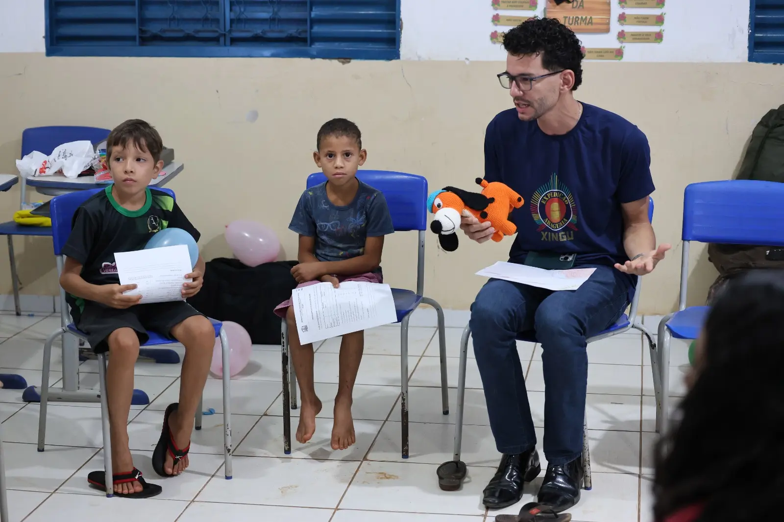 Momento de partilha no Círculo de Paz: educador fala com as crianças mostrando o brinquedo mascote. Ao lado, meninos observam atentos com folhas e balões