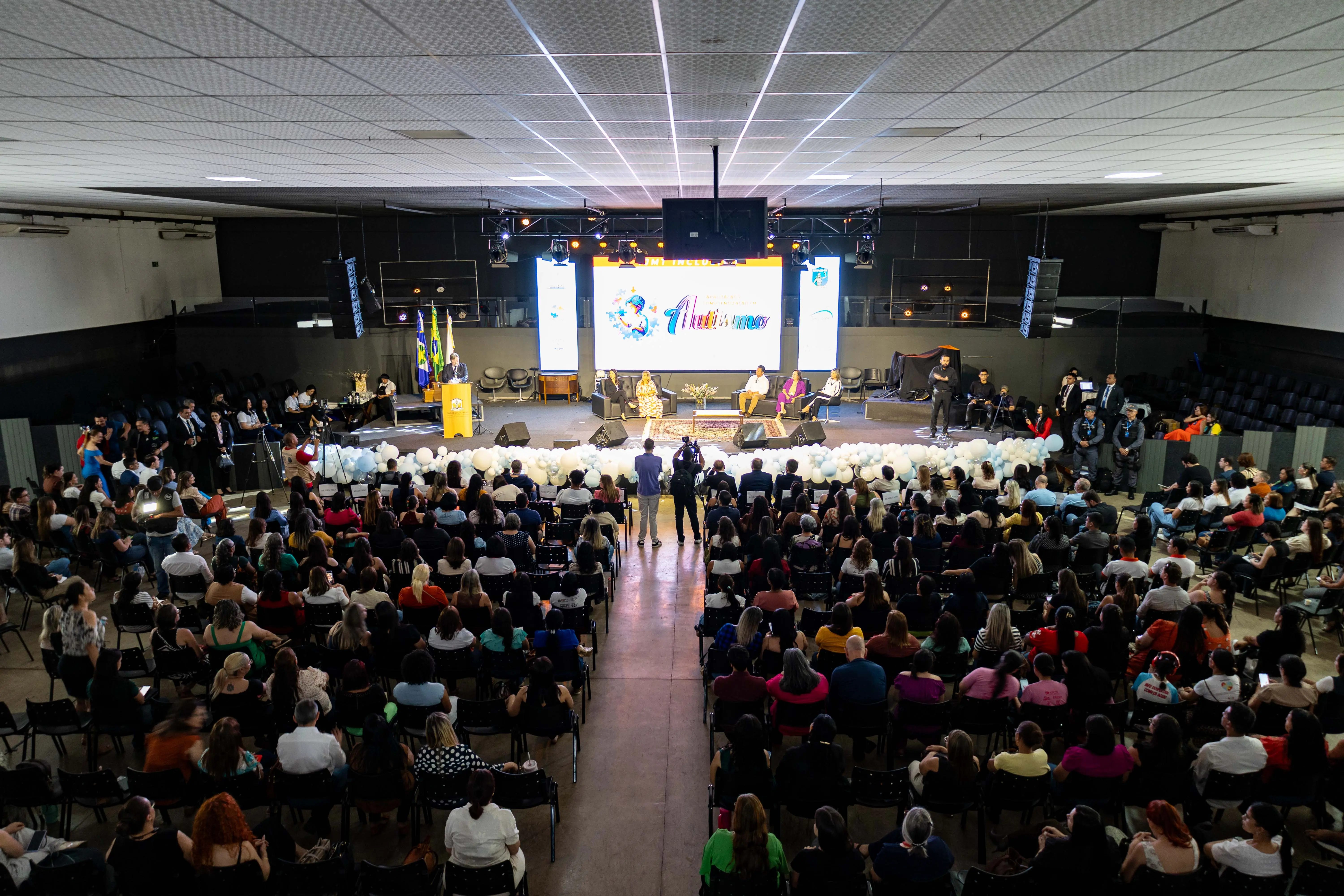 Imagem aérea mostra auditório lotado em Rondonópolis durante o evento TJMT Inclusivo. No palco, representantes do Judiciário e parceiros falam sobre autismo diante de grande público.