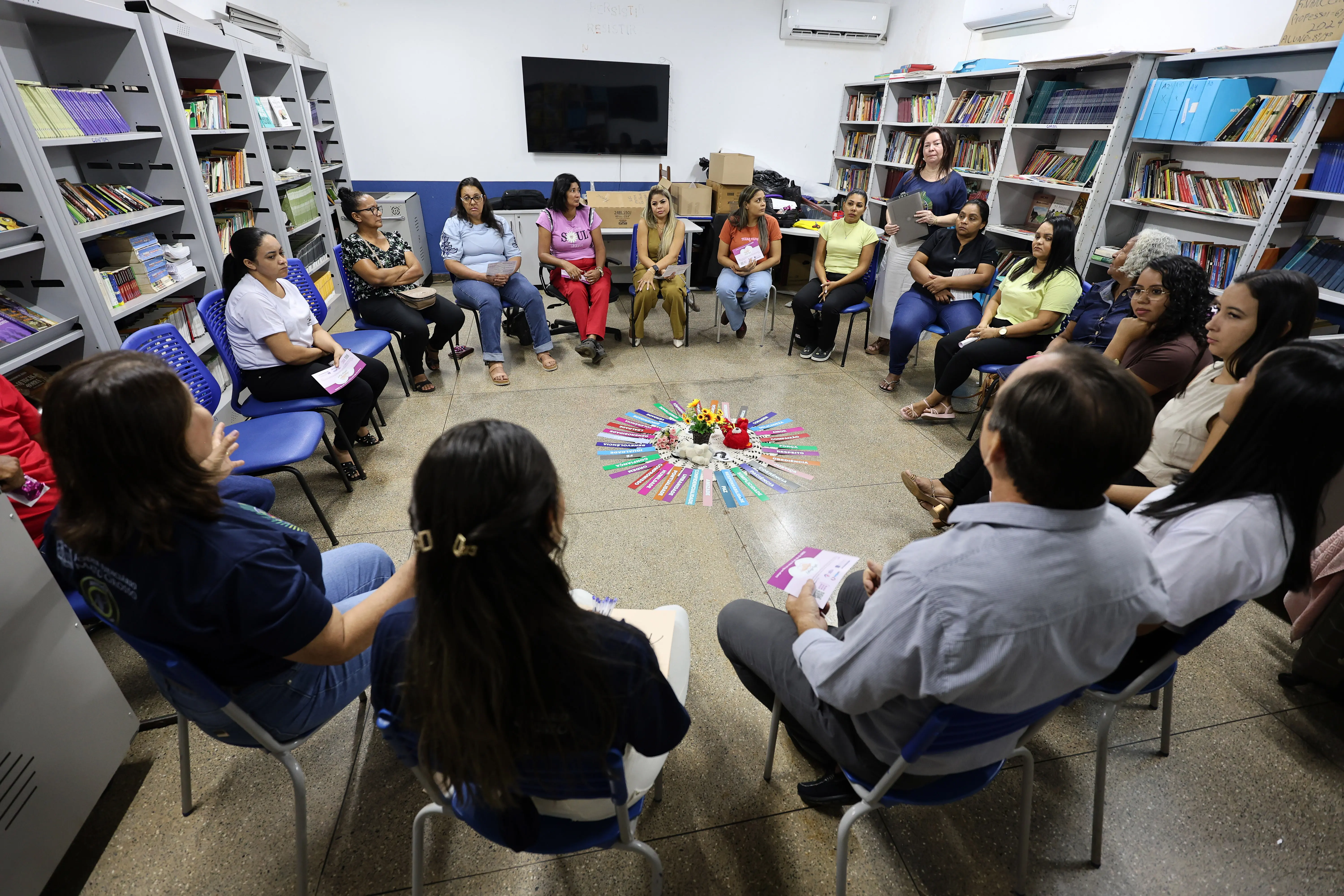 Grupo sentado em círculo numa sala com estantes repletas de livros. No centro, há um arranjo colorido no chão, sugerindo dinâmica coletiva. O ambiente é acolhedor, típico de biblioteca comunitária.